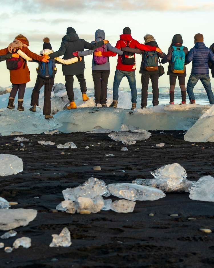 A WeRoad group trip with their arms around each other, standing on a large block of ice on a black sand beach.