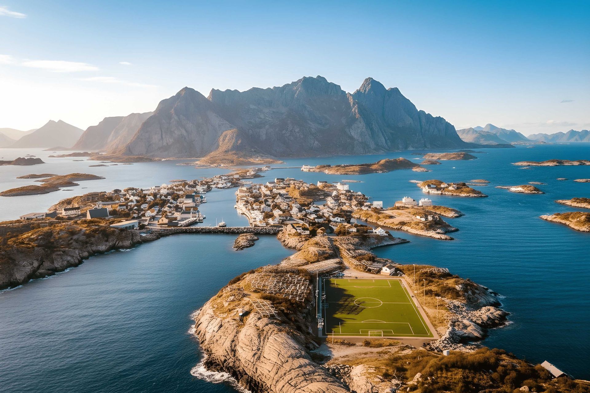 Eine Luftaufnahme eines Fußballplatzes auf einer felsigen Halbinsel neben einem Küstendorf, umgeben von Inseln und Bergen.