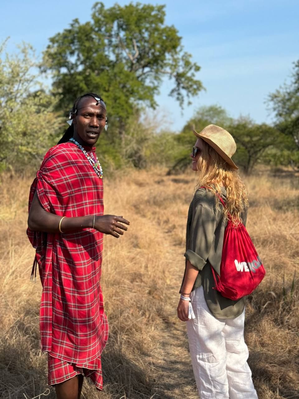 A Maasai man in traditional red attire talking to a woman from a WeRoad group trip in a grassy savanna.