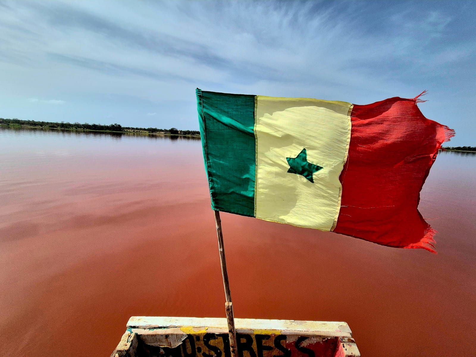 La bandera senegalesa, izada en la proa de un barco, ondea al viento sobre un gran lago rosa bajo un cielo azul con nubes.