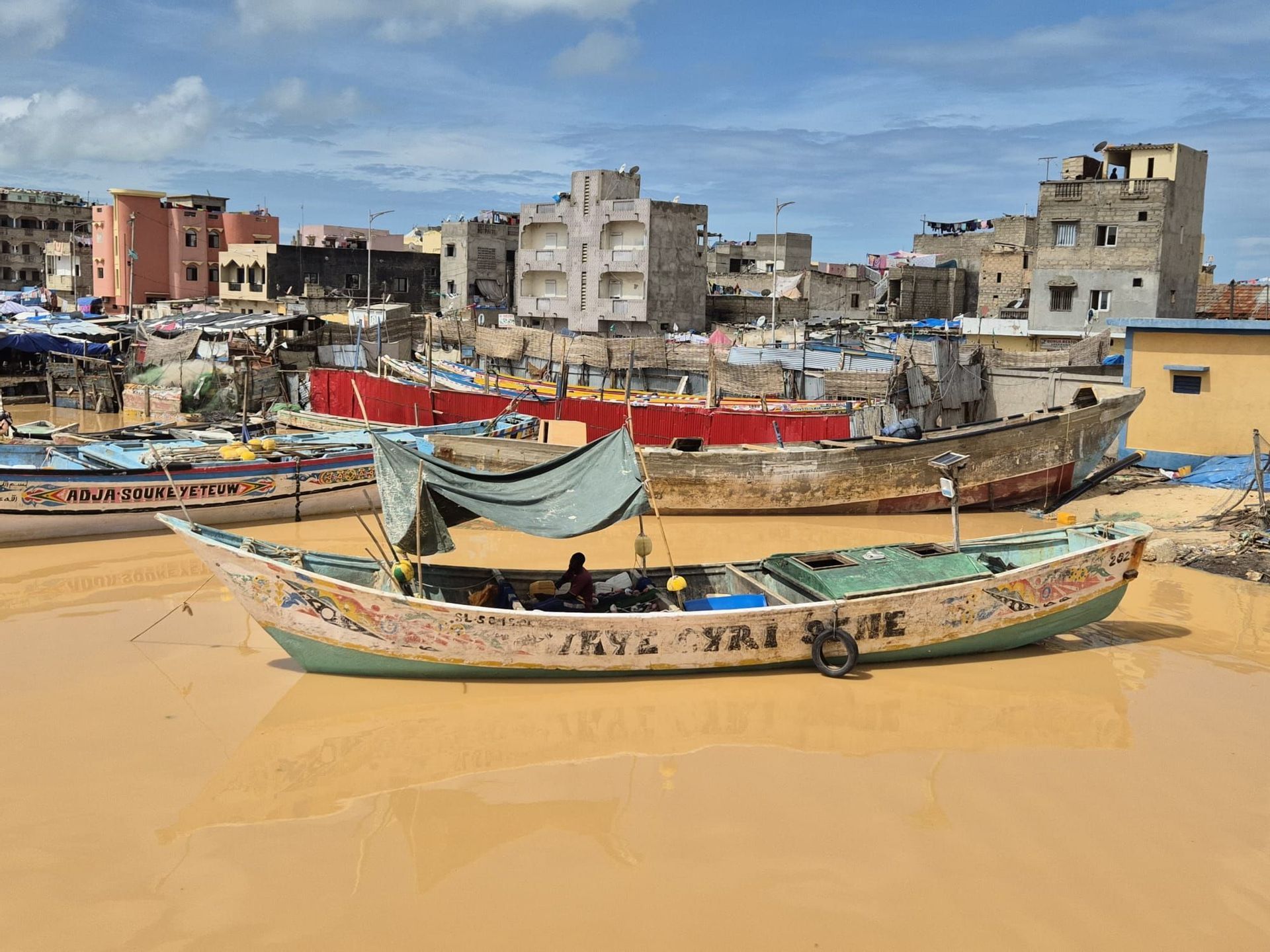 Una colorida barca de pesca con un toldo de lona flota en agua fangosa en un puerto abarrotado, con un paisaje urbano de fondo.