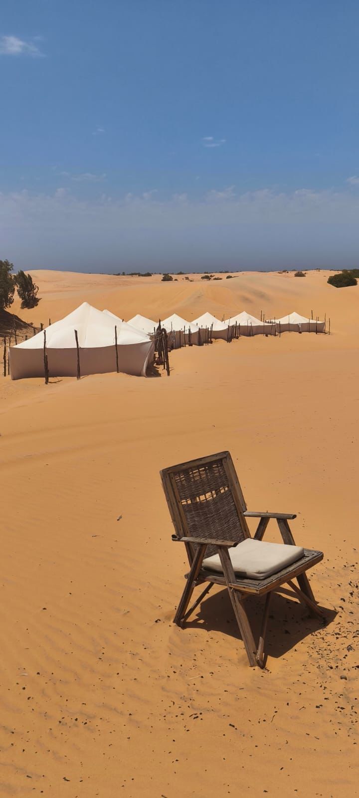 Una silla de madera se sitúa en primer plano sobre una duna de arena anaranjada, con una fila de tiendas blancas formando un campamento en el desierto al fondo.