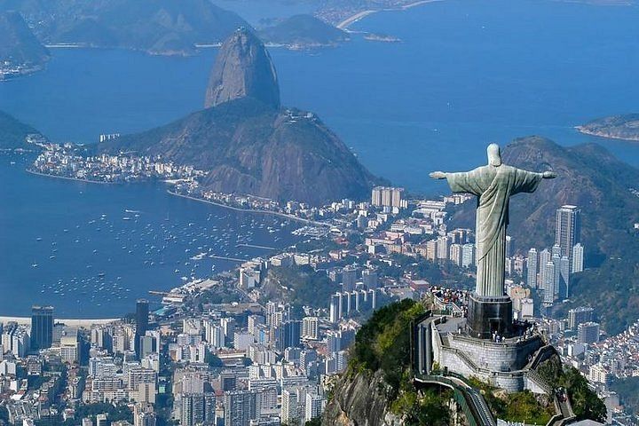 Una grande statua con le braccia aperte su una cima di montagna, che domina una vasta città costiera e una grande baia con un'altra montagna.