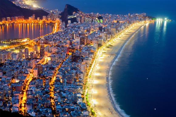 Vista aerea notturna di una città costiera splendidamente illuminata, con una lunga spiaggia risplendente che si curva lungo l'oceano scuro.