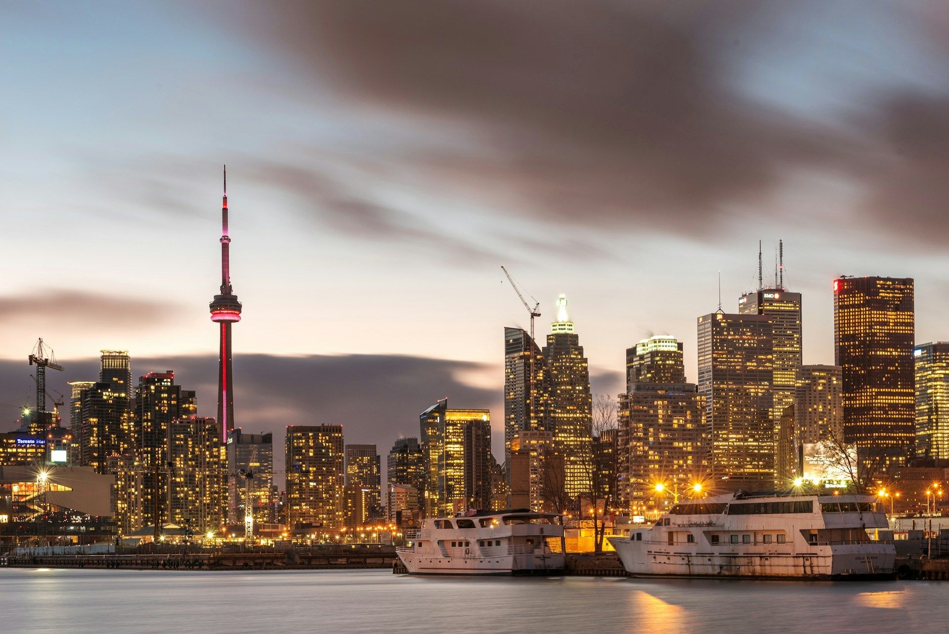 A city skyline filled with illuminated skyscrapers and a tall, red-lit tower at dusk, viewed from across the water where two boats are docked.