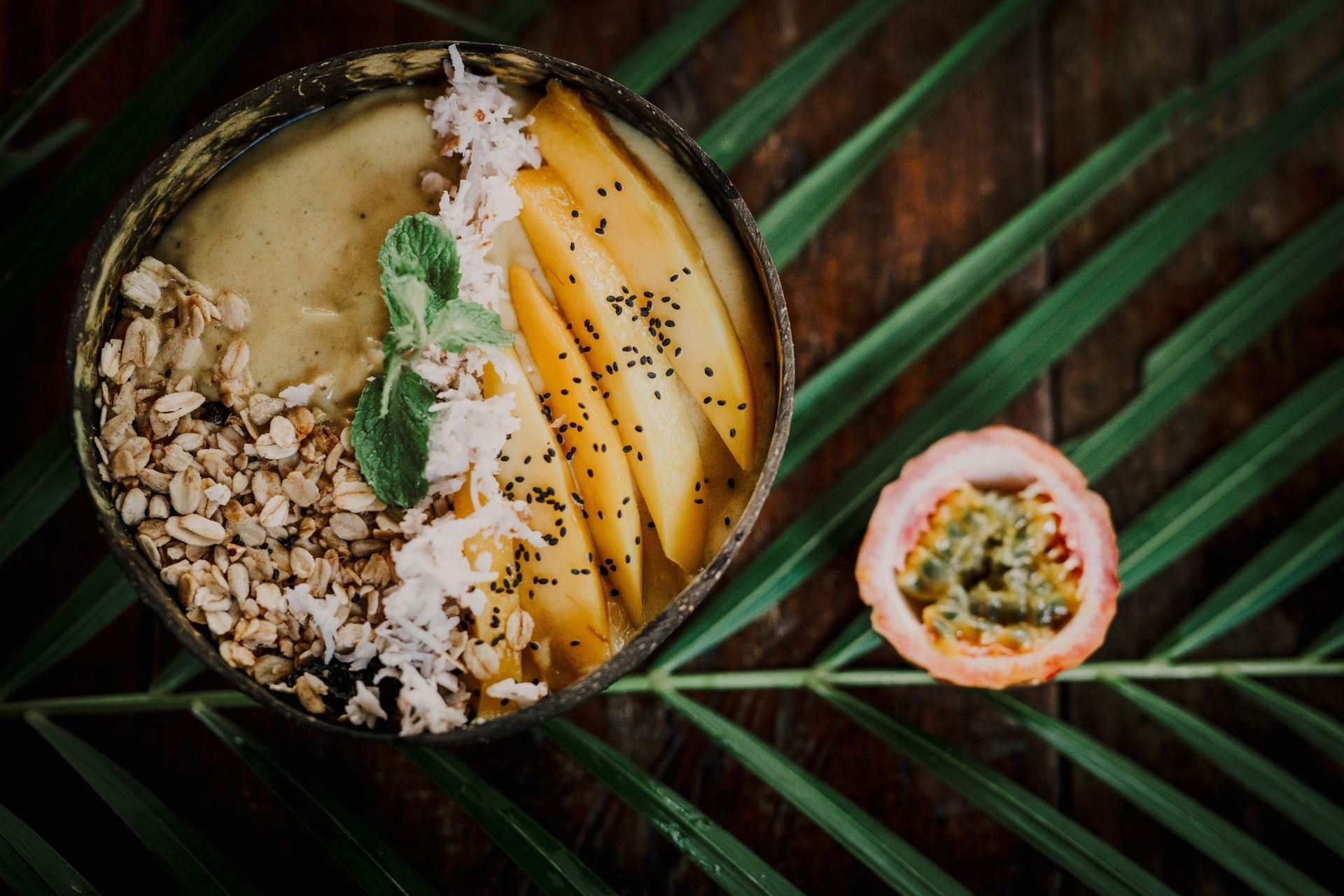 Un bol de smoothie mangue, granola et copeaux de coco dans une coque de noix de coco, près d'un fruit de la passion sur une feuille de palmier.