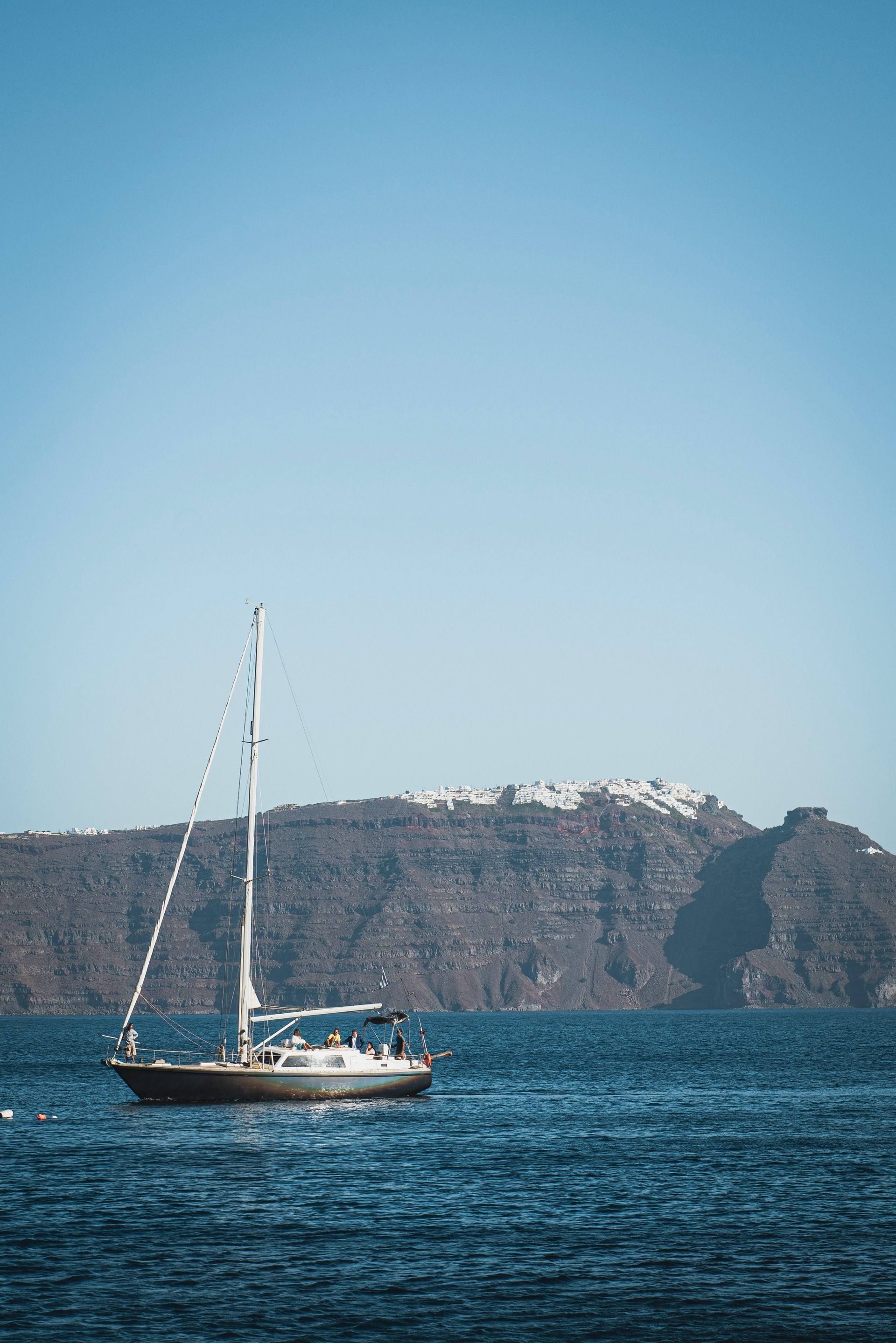 Una barca a vela con un gruppo WeRoad naviga sul mare blu di fronte a un'imponente scogliera con edifici bianchi in cima.