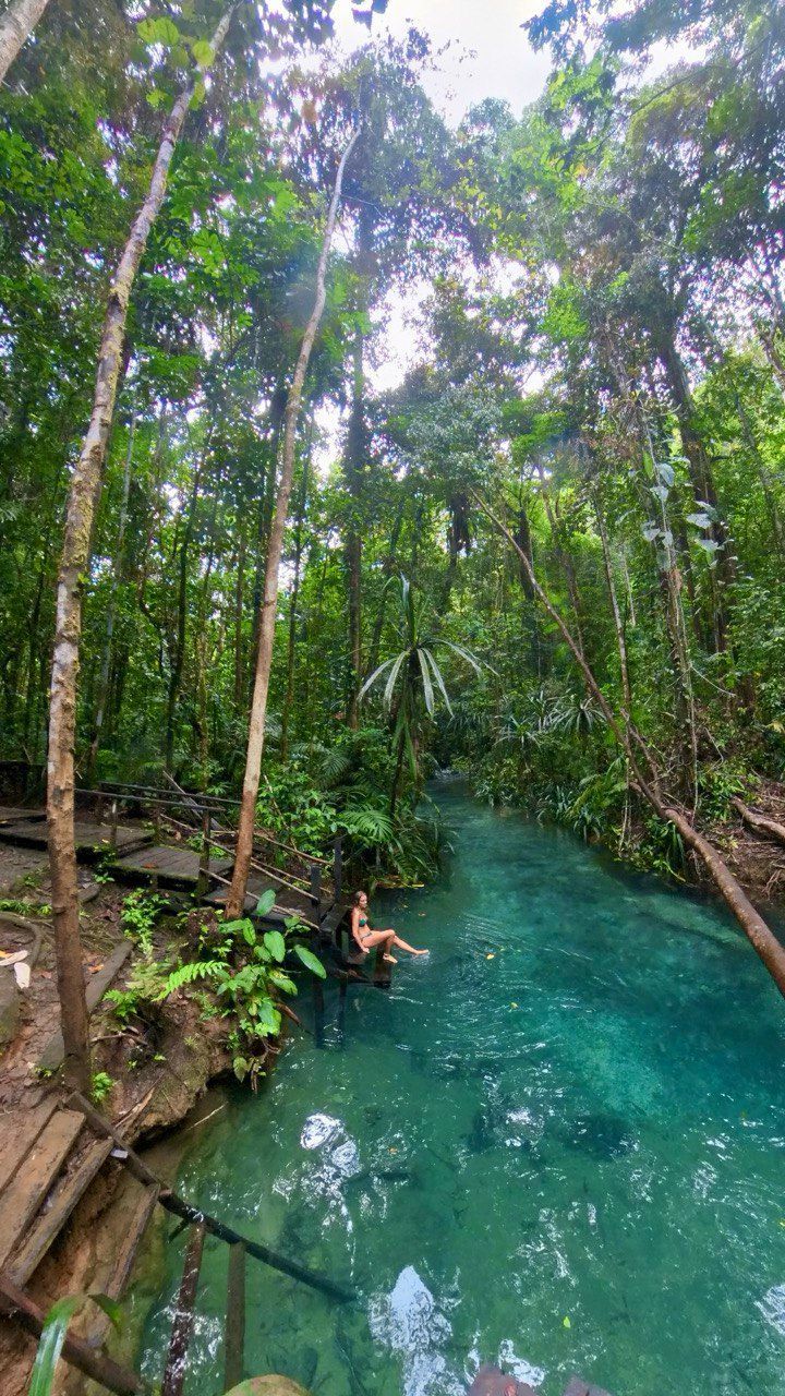 Una donna in bikini siede su una piattaforma di legno, immergendo i piedi in un limpido fiume turchese che attraversa una giungla fitta.