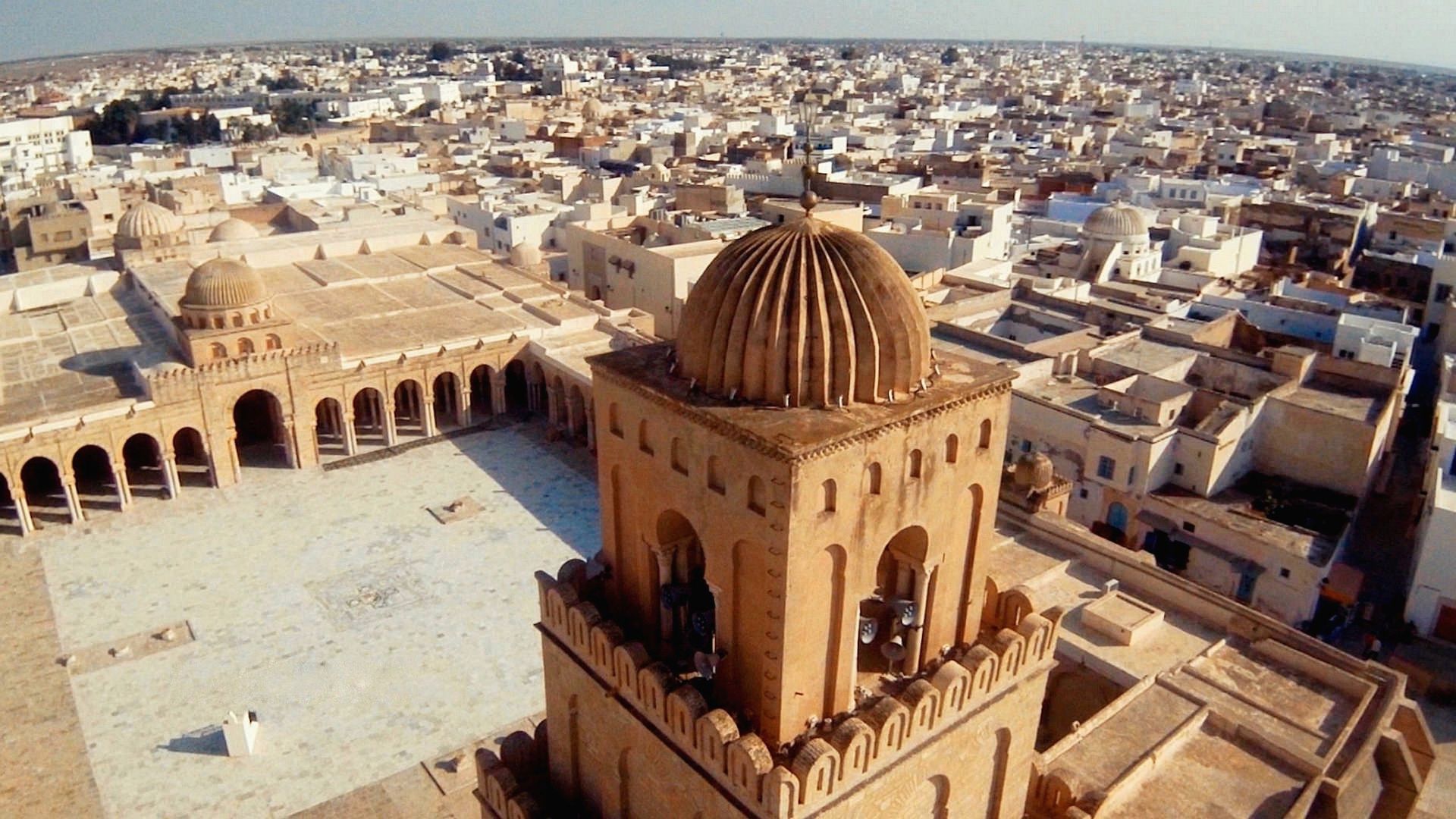 Una vista aerea di una grande moschea con una torre a cupola e un ampio cortile, circondata da una città densamente popolata sotto un cielo sereno.