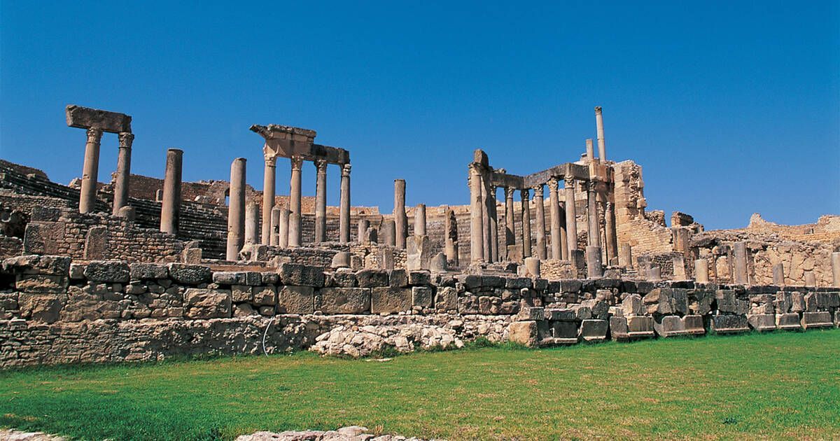 D'anciennes ruines de pierre, avec des rangées de colonnes, se dressent sur un champ herbeux sous un ciel bleu clair.