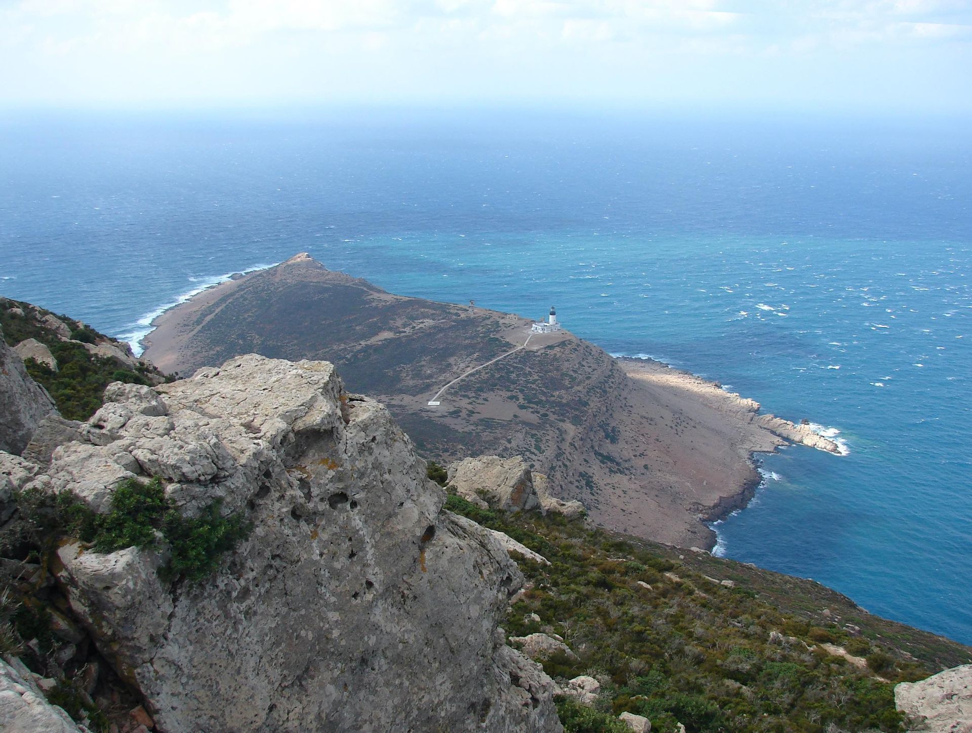 Vue d'une falaise rocheuse dominant une péninsule avec un phare blanc, entourée par la mer bleue.