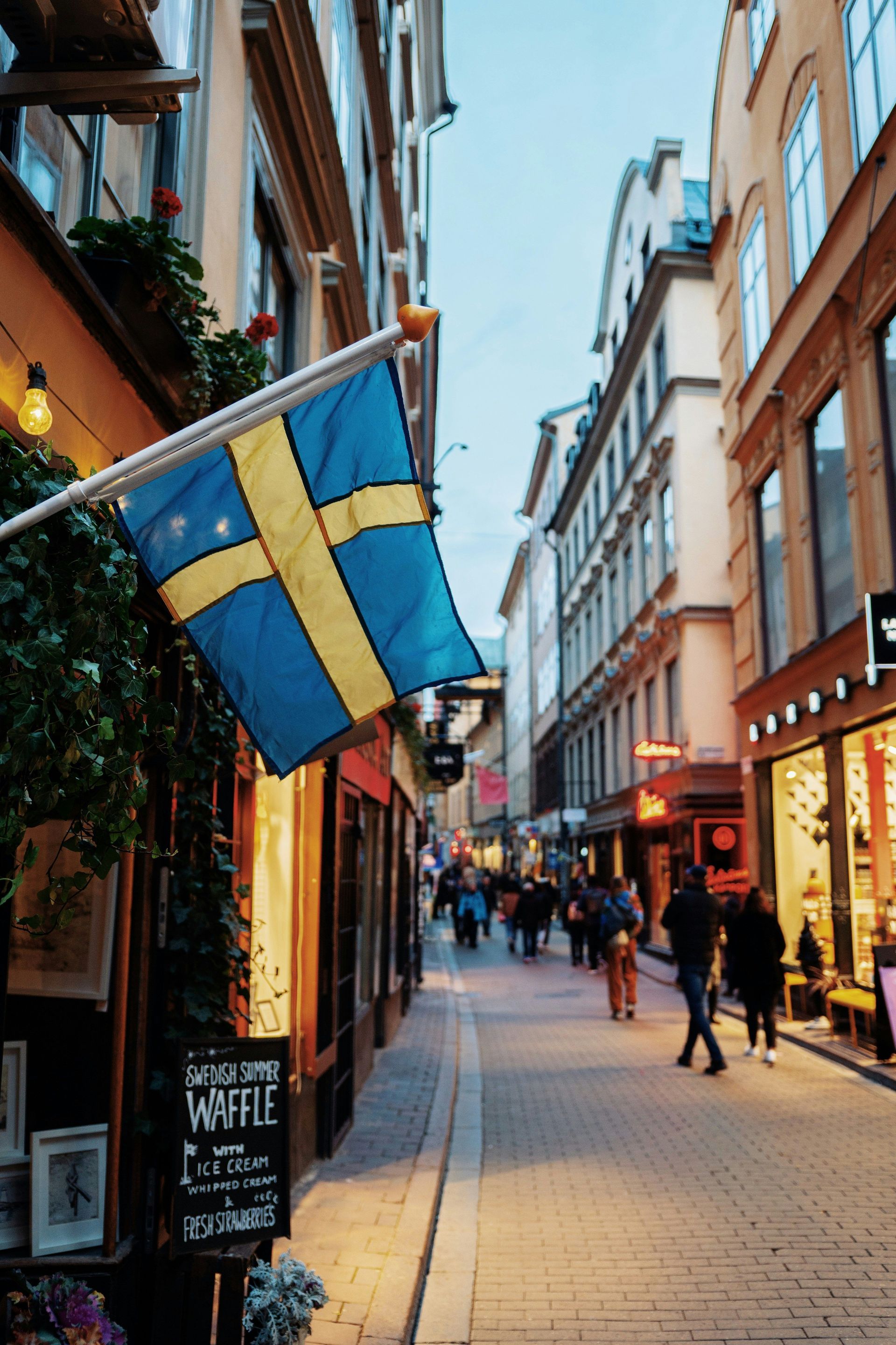 A Swedish flag hangs from a pole on a building along a narrow cobblestone street filled with shops and people.