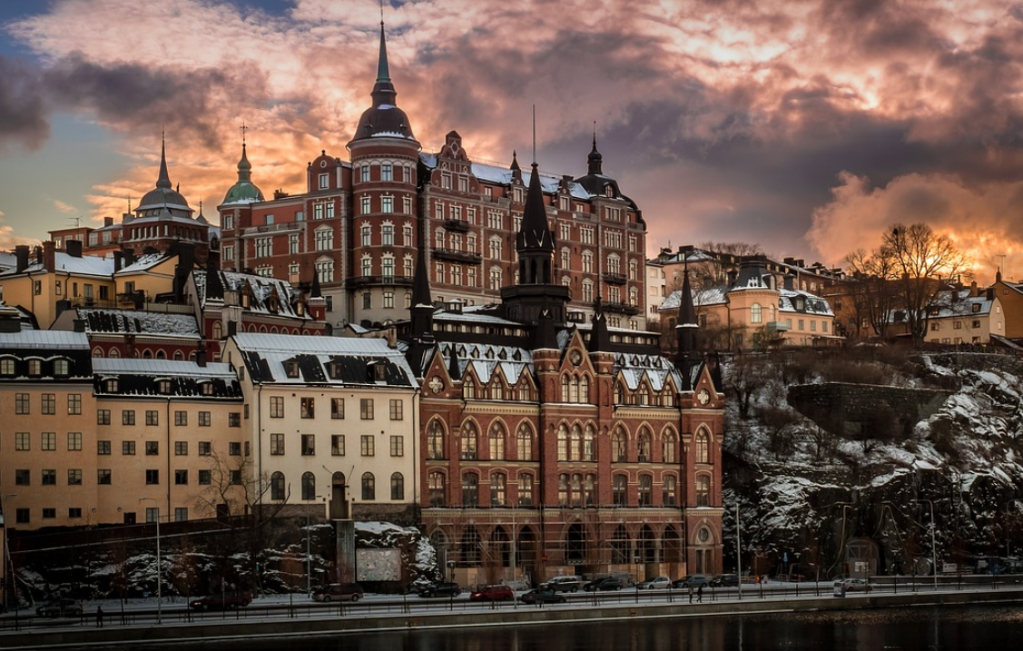 Ornate city buildings with snow-covered roofs stand on a rocky hillside under a dramatic sunset sky.