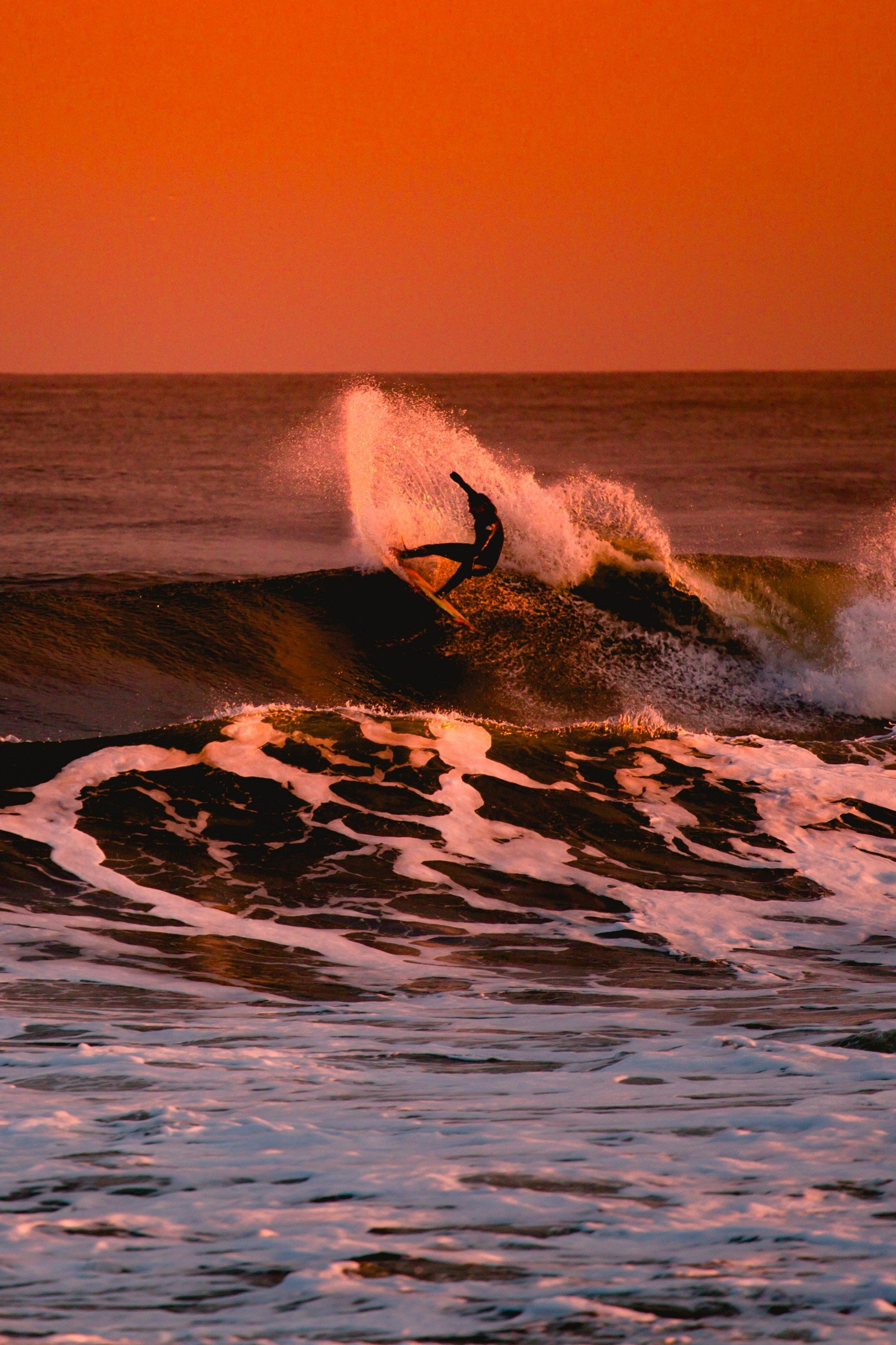 Un surfista in silhouette cavalca una grande onda oceanica, sollevando spruzzi contro un cielo arancione brillante al tramonto.