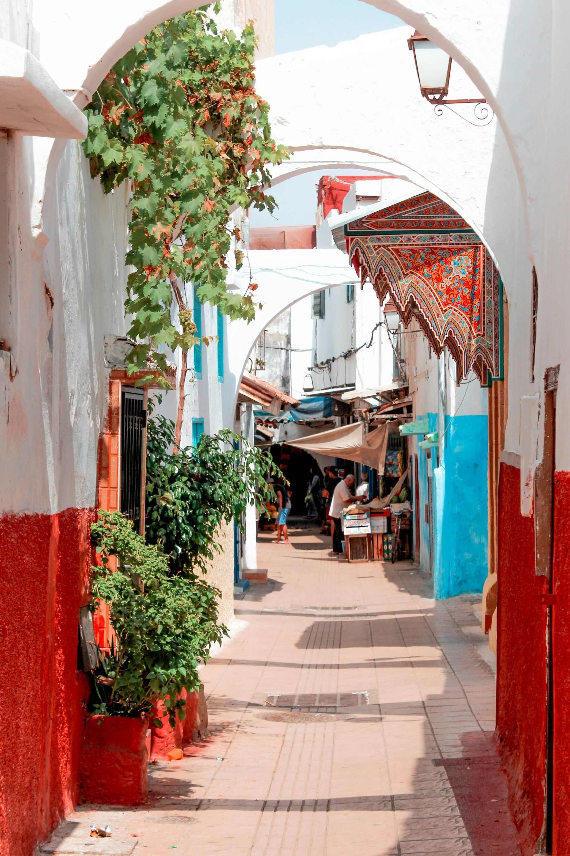 Una vista de un callejón estrecho y soleado con arcos blancos, paredes rojas y azules vibrantes, y puestos de mercado a lo lejos.