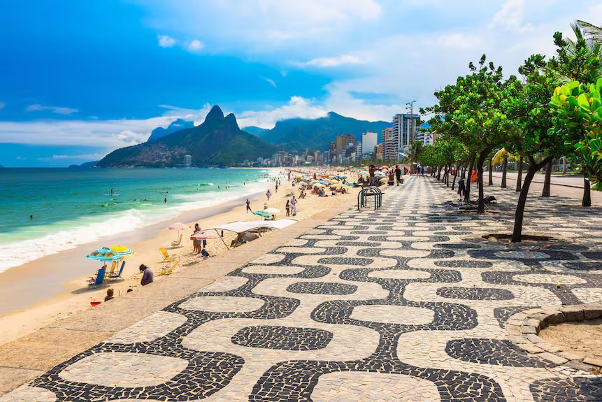 Una spiaggia soleggiata e affollata accanto a una città, con un caratteristico lungomare a motivi bianchi e neri e grandi montagne visibili in lontananza.