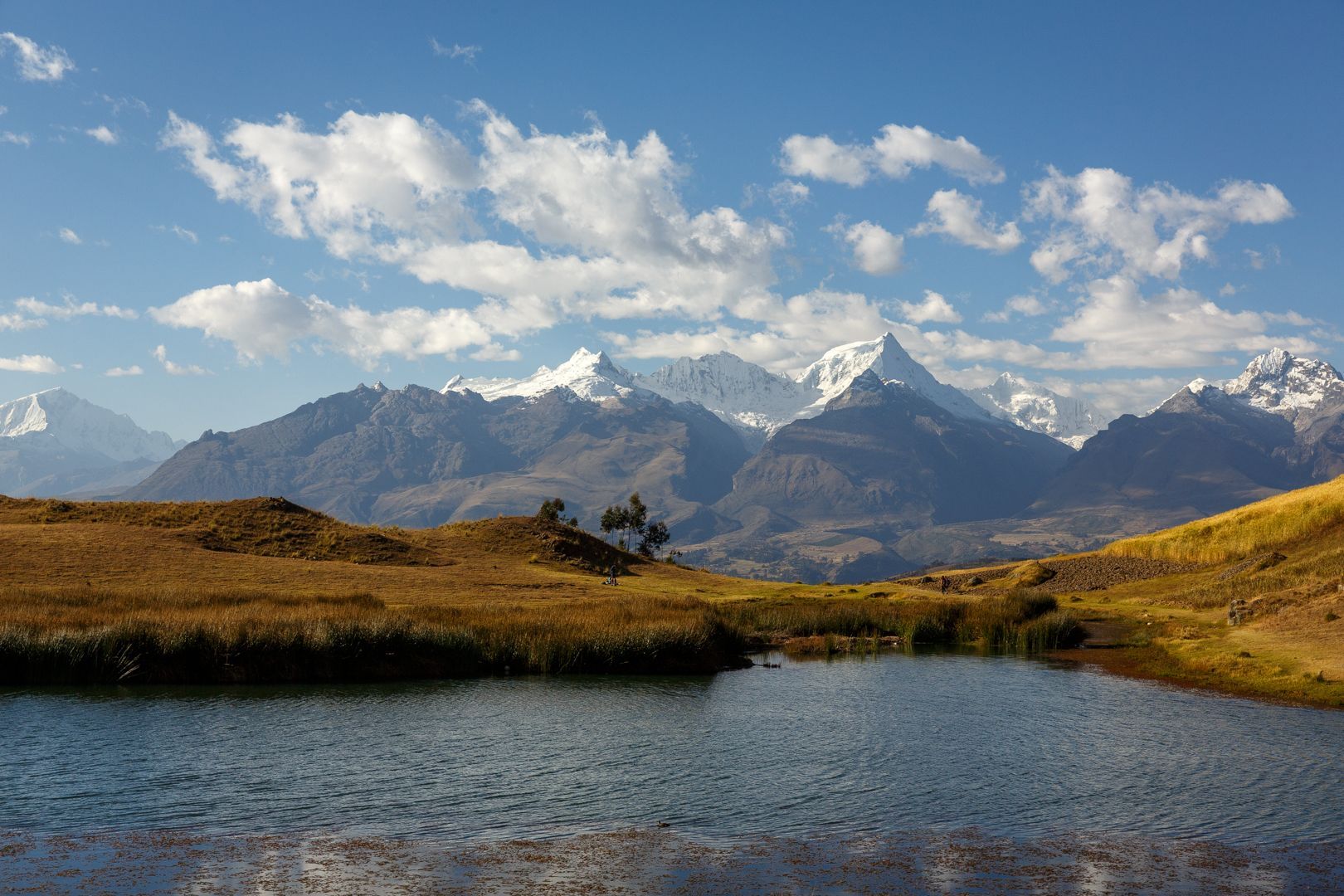 Un lago di montagna si trova in una valle erbosa di fronte a una lontana catena montuosa innevata sotto un cielo parzialmente nuvoloso.
