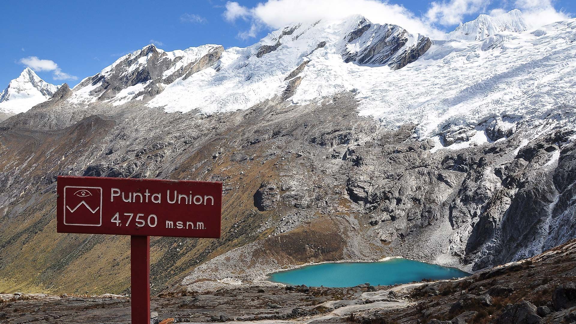 Un segnavia rosso si erge in un paesaggio roccioso con un lago alpino turchese sotto montagne innevate.