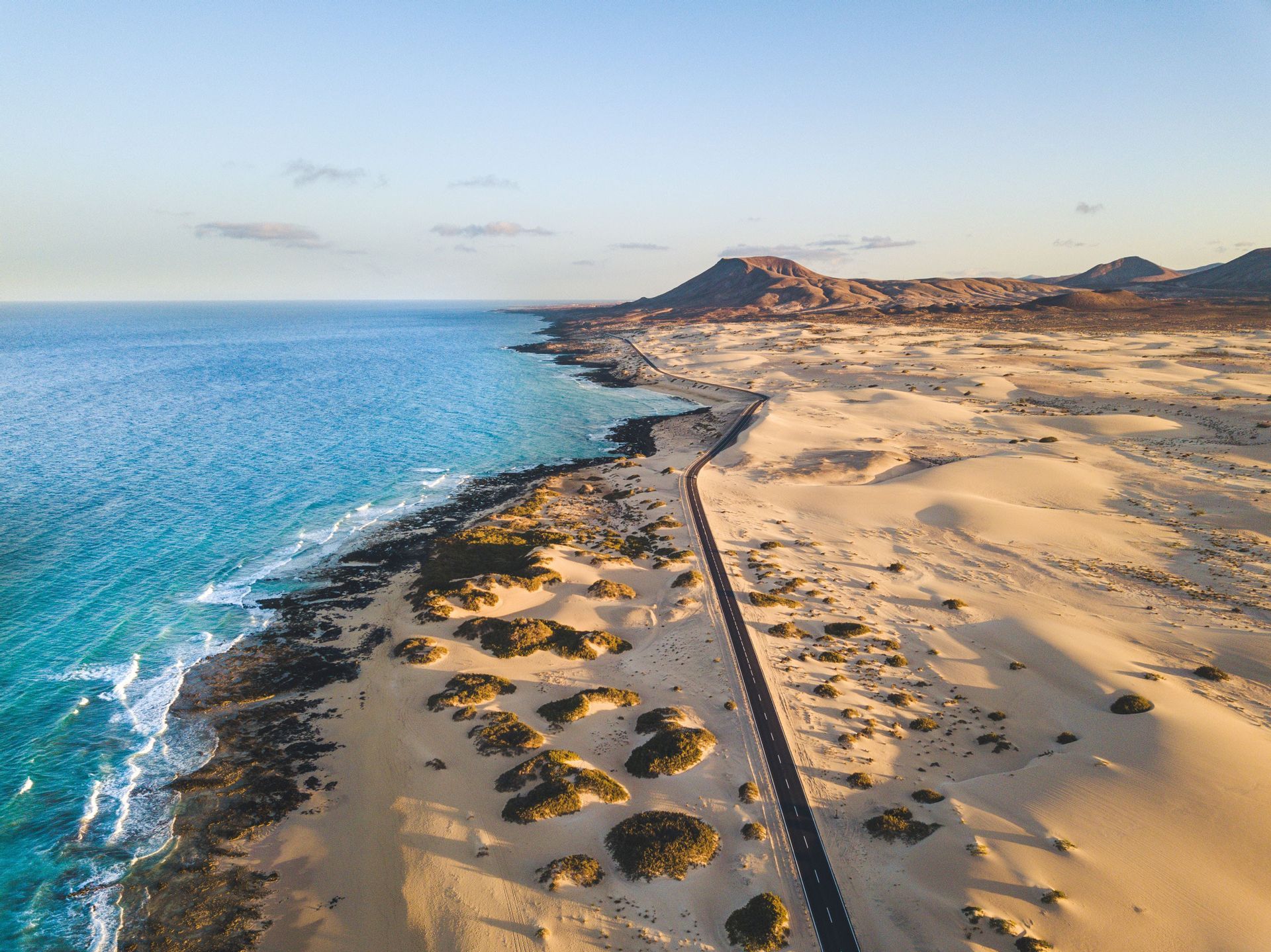 Una veduta aerea di una lunga strada che si snoda tra vaste dune di sabbia e una costa oceanica turchese, con montagne in lontananza.