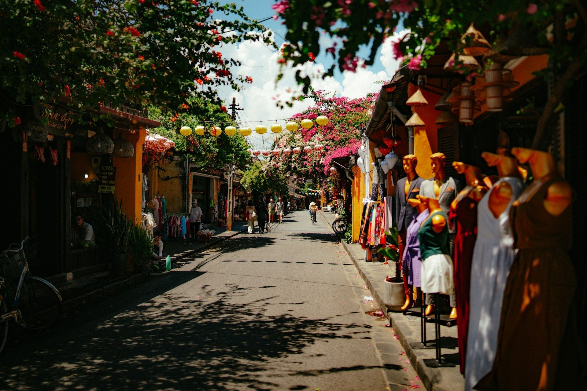 Una strada assolata fiancheggiata da negozi colorati, manichini e alberi fioriti, con lanterne gialle appese in alto.
