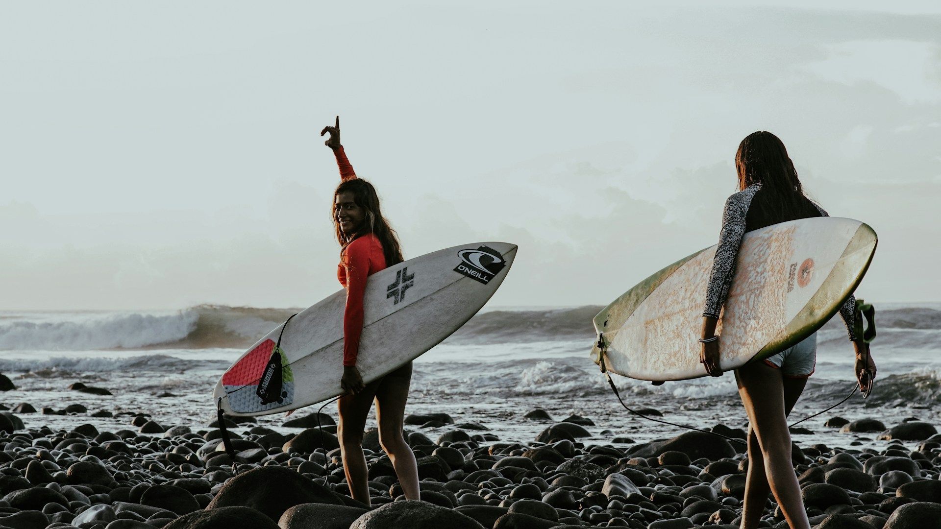 Due donne di un viaggio di gruppo WeRoad trasportano le loro tavole da surf su una spiaggia rocciosa, con le onde dell'oceano sullo sfondo.