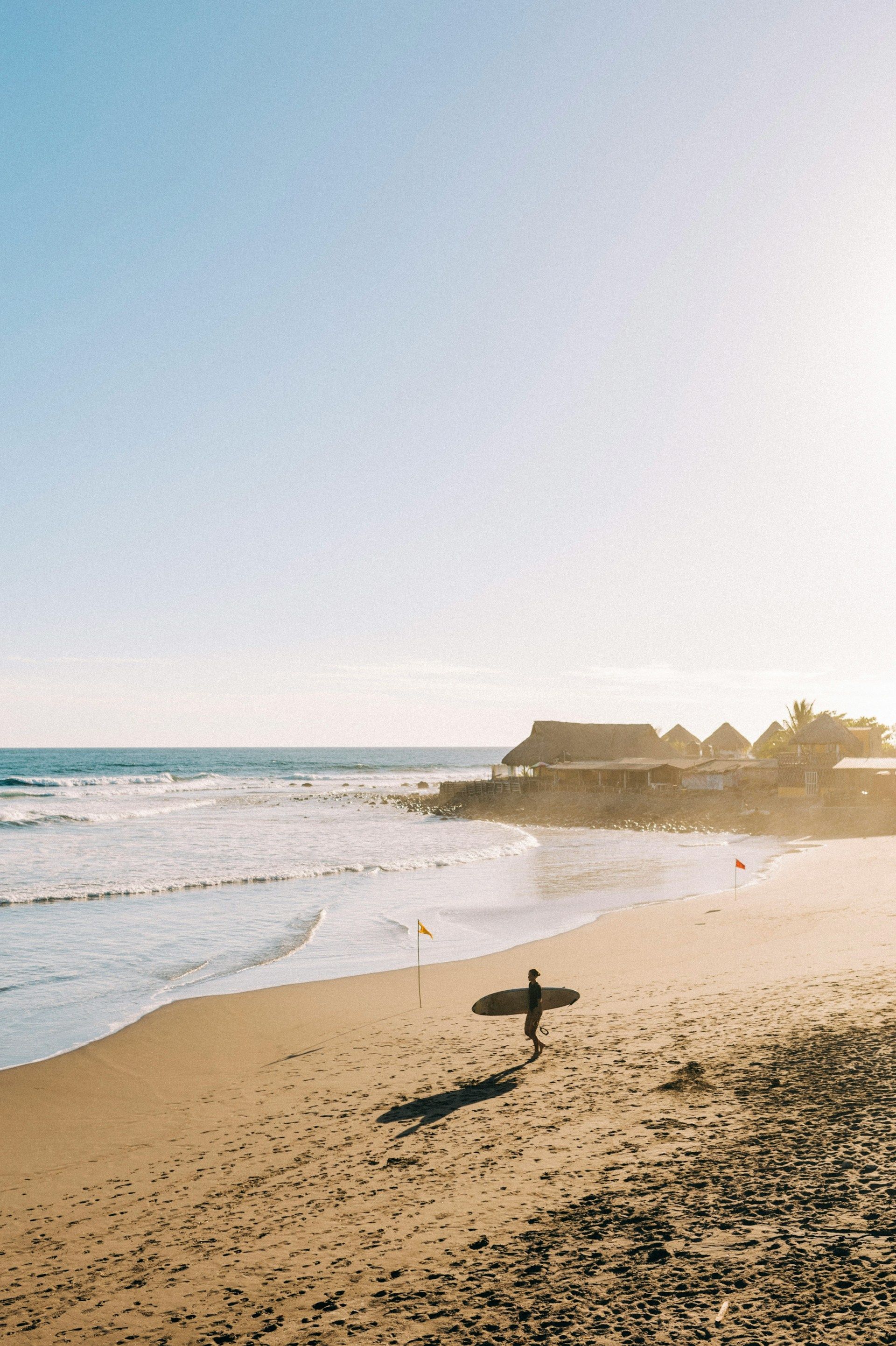 Un surfista in controluce cammina su una spiaggia sabbiosa con la sua tavola da surf, sullo sfondo l'oceano e le capanne balneari.