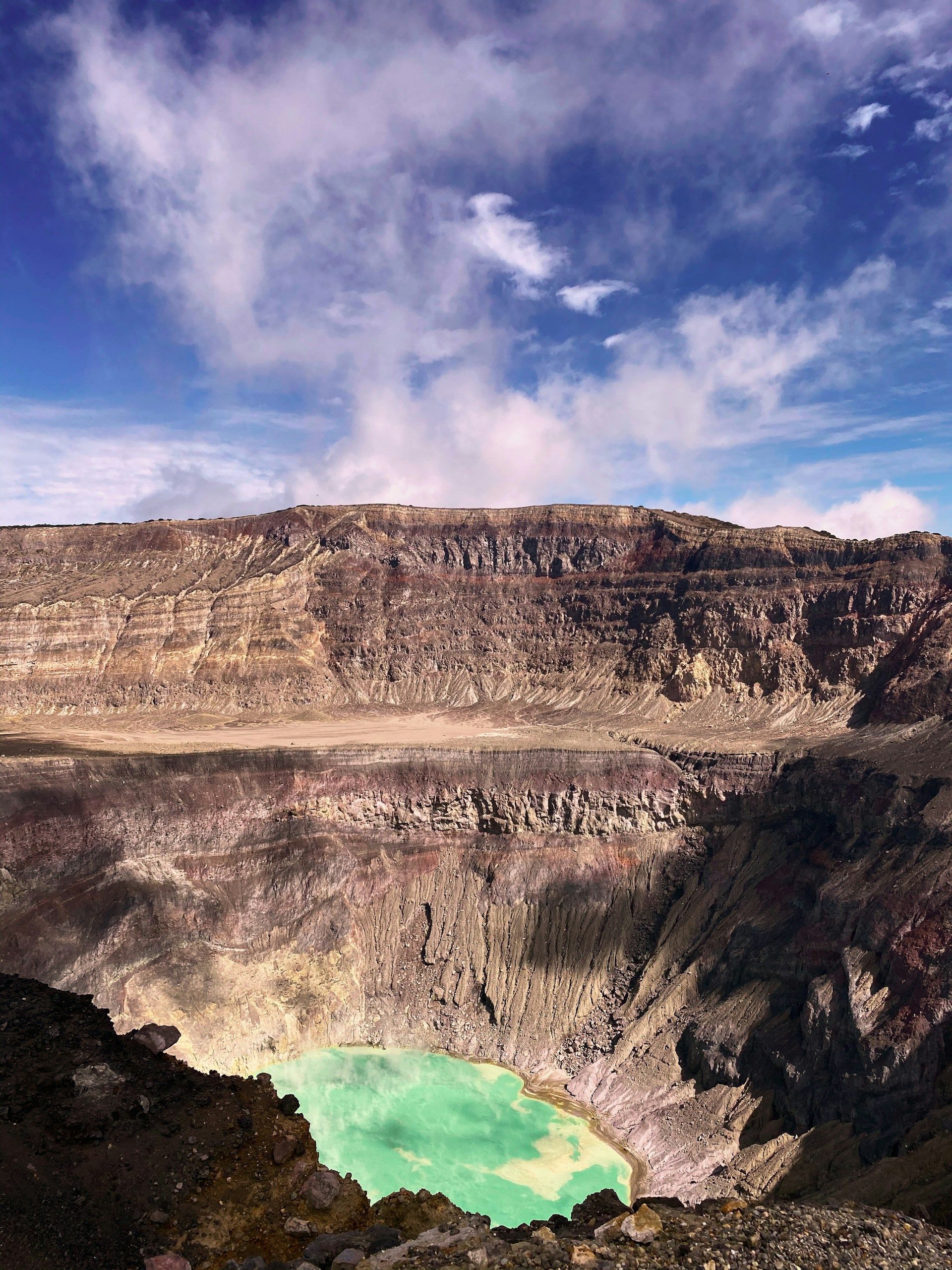 Una vista su un vasto cratere vulcanico con pareti rocciose stratificate e un lago turchese sul fondo, sotto un cielo blu con nuvole bianche.
