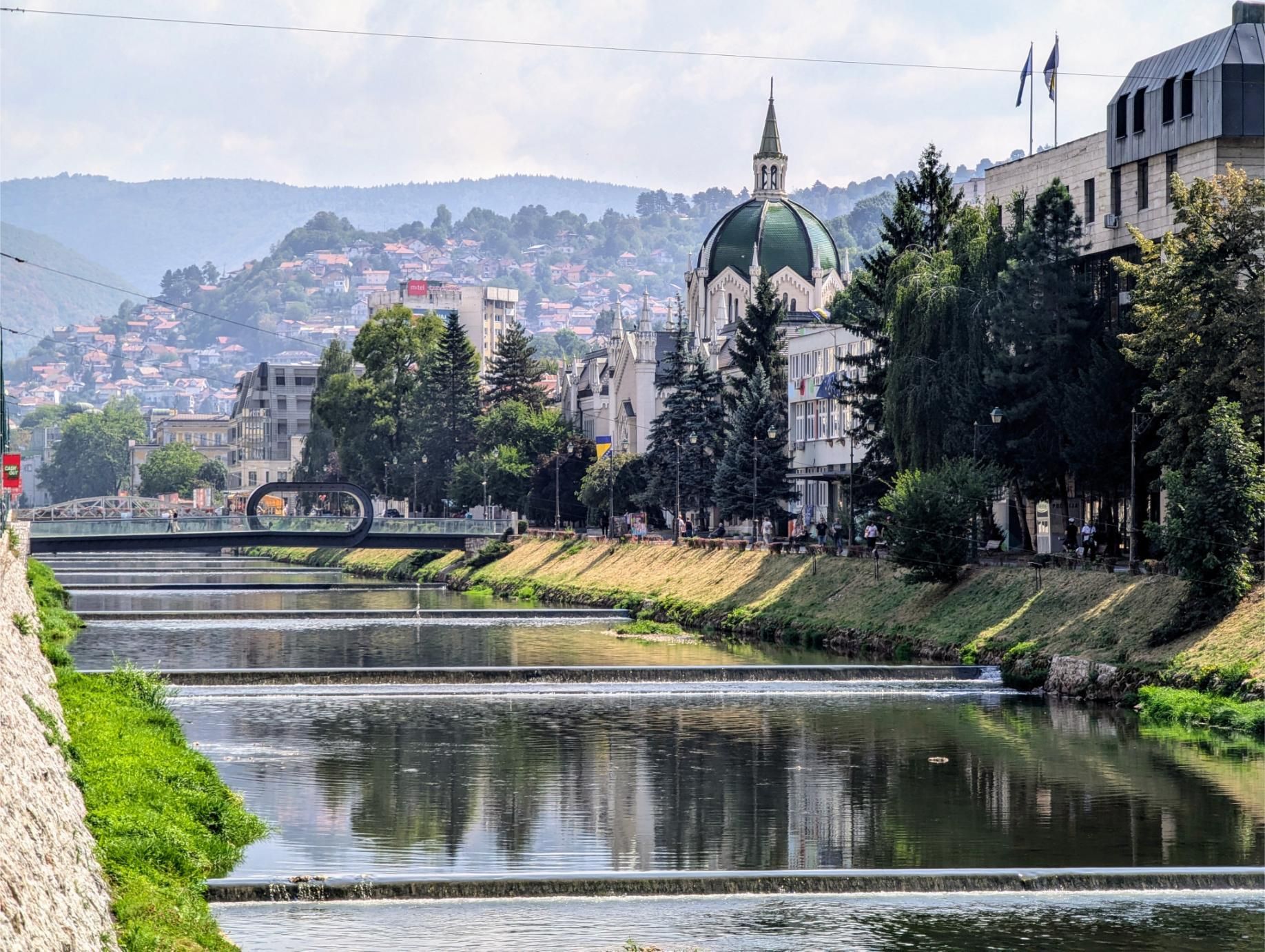 Un fiume scorre su piccoli sbarramenti attraverso una città con un edificio dalla cupola verde e una collina ricoperta di case.