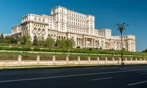 Un imponente edificio neoclassico bianco si erge su un pendio erboso dietro una recinzione in pietra, visto dall'altra parte di una strada vuota.