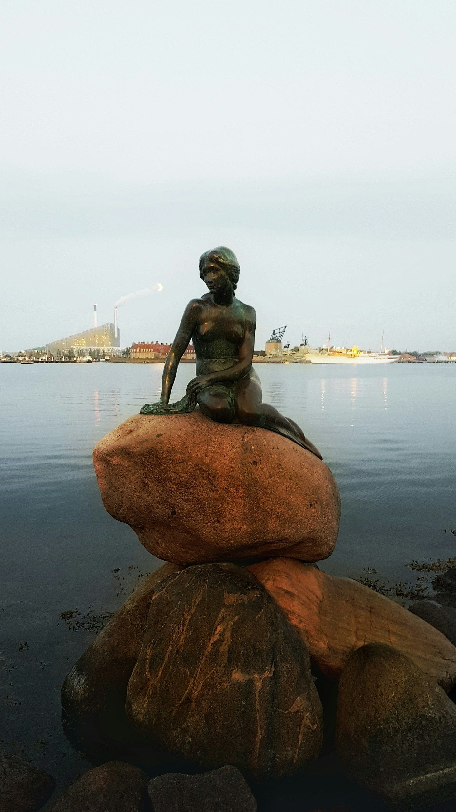 Une statue de sirène en bronze assise sur un grand rocher au bord de l'eau, avec la silhouette d'une ville visible en arrière-plan au crépuscule.
