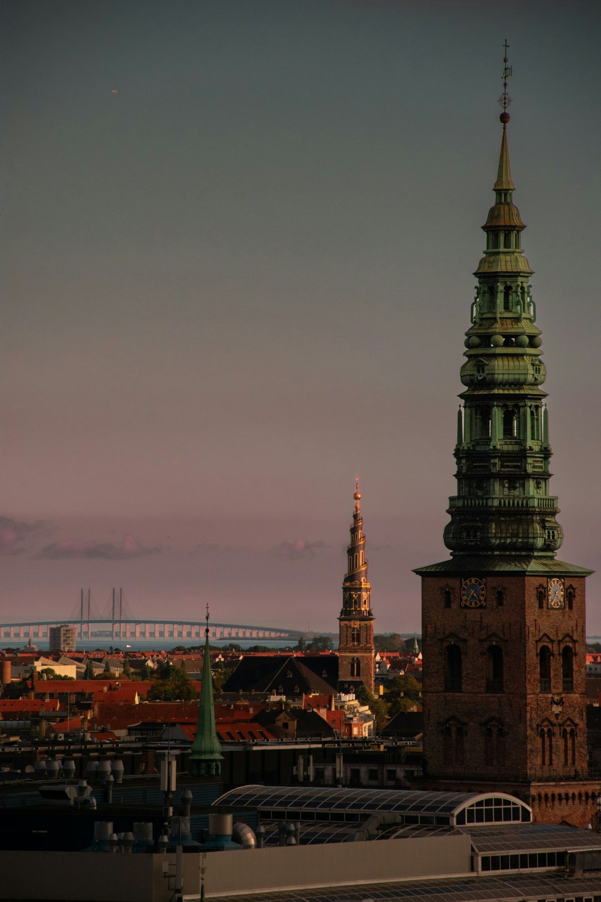 Veduta della città al tramonto con ornate torri di chiesa e un ponte sospeso in lontananza.