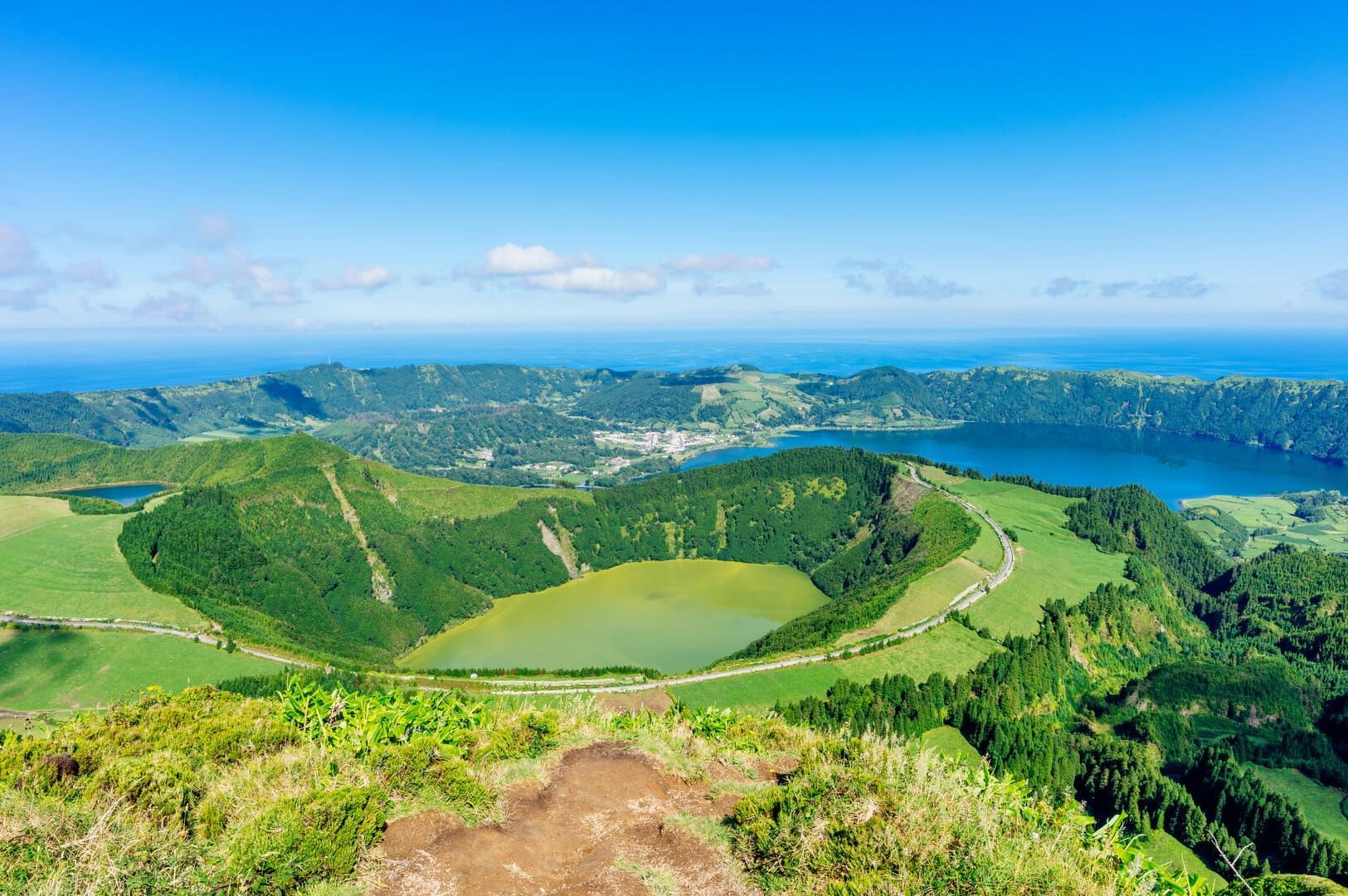 An aerial view of green volcanic hills surrounding two crater lakes, one green and one blue, with the ocean on the horizon.