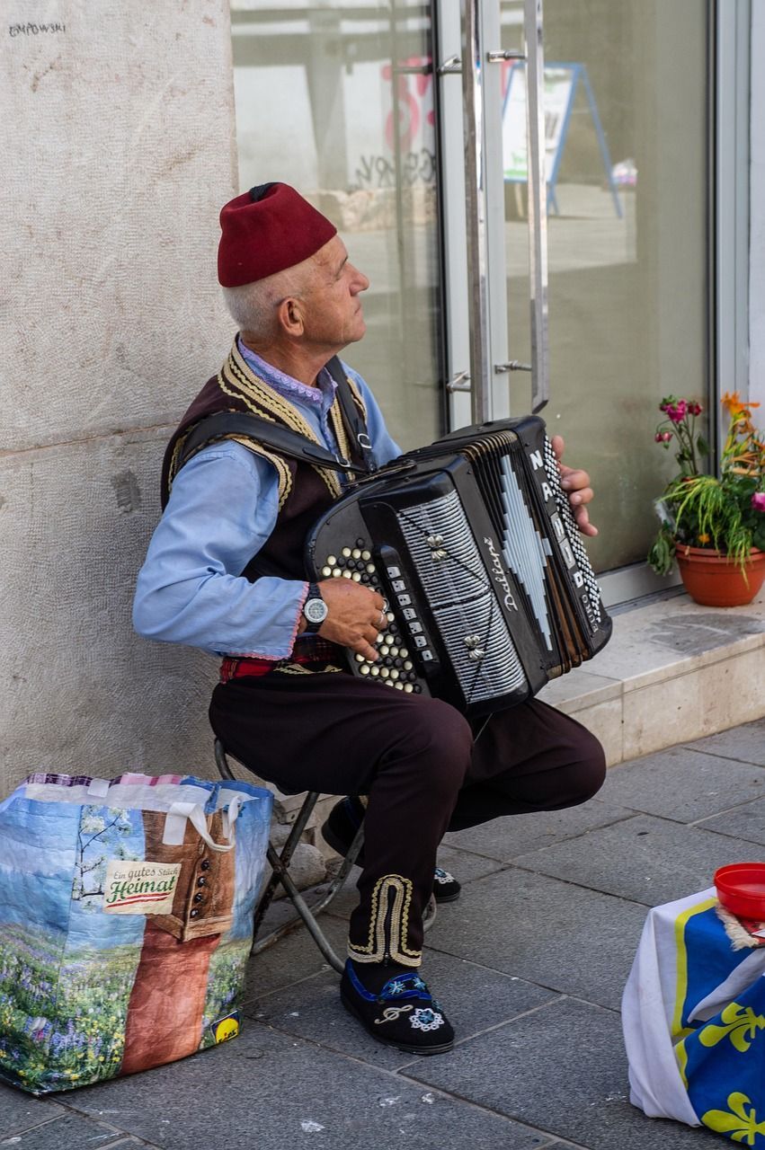 Un uomo con un fez rosso e un gilet tradizionale suona una fisarmonica nera mentre è seduto su uno sgabello su un marciapiede.