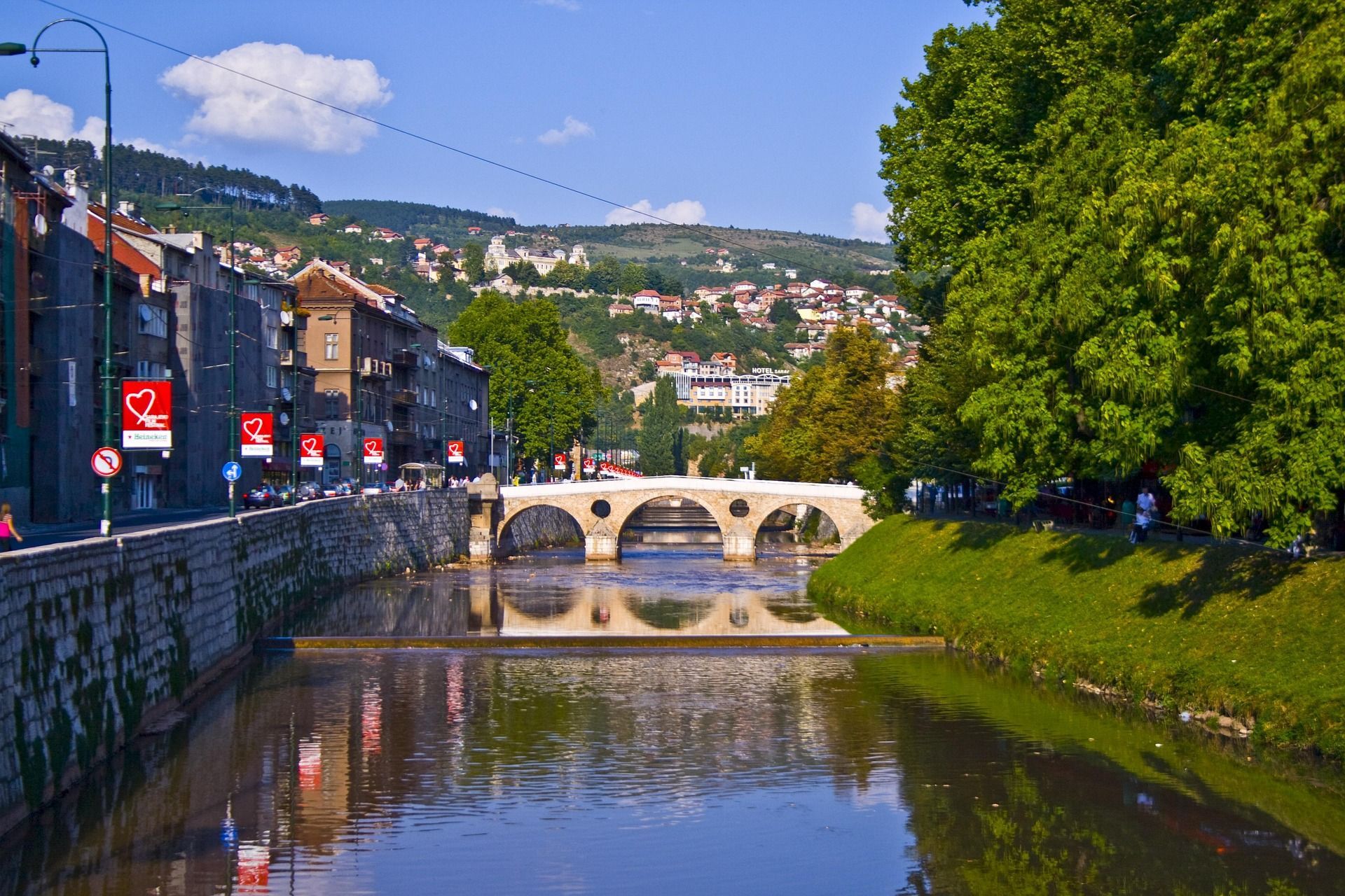 Un ponte in pietra ad arco attraversa un fiume che scorre in una città con edifici e una riva erbosa e alberata sotto un cielo azzurro e limpido.