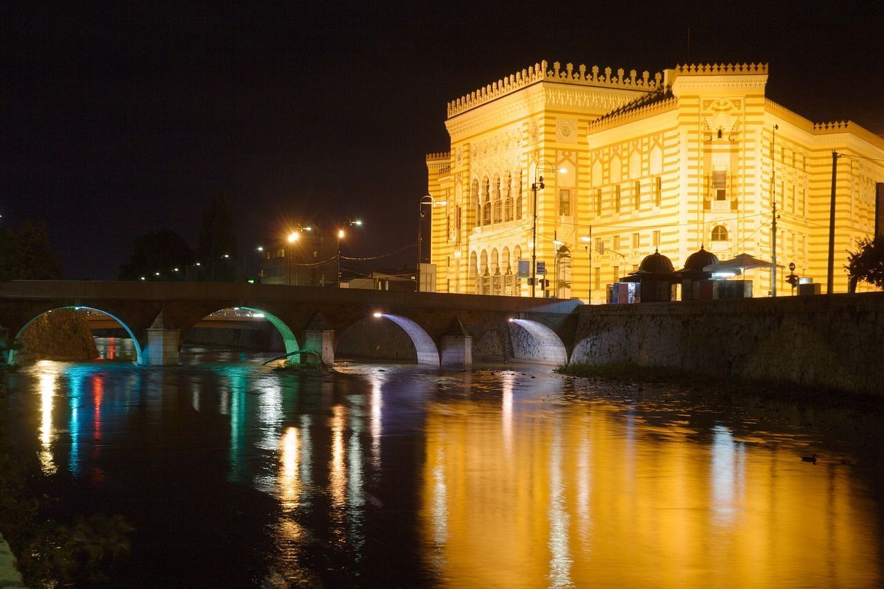 Un sontuoso edificio e un ponte ad arco in pietra sono illuminati di notte, le loro luci si riflettono su un ampio fiume.