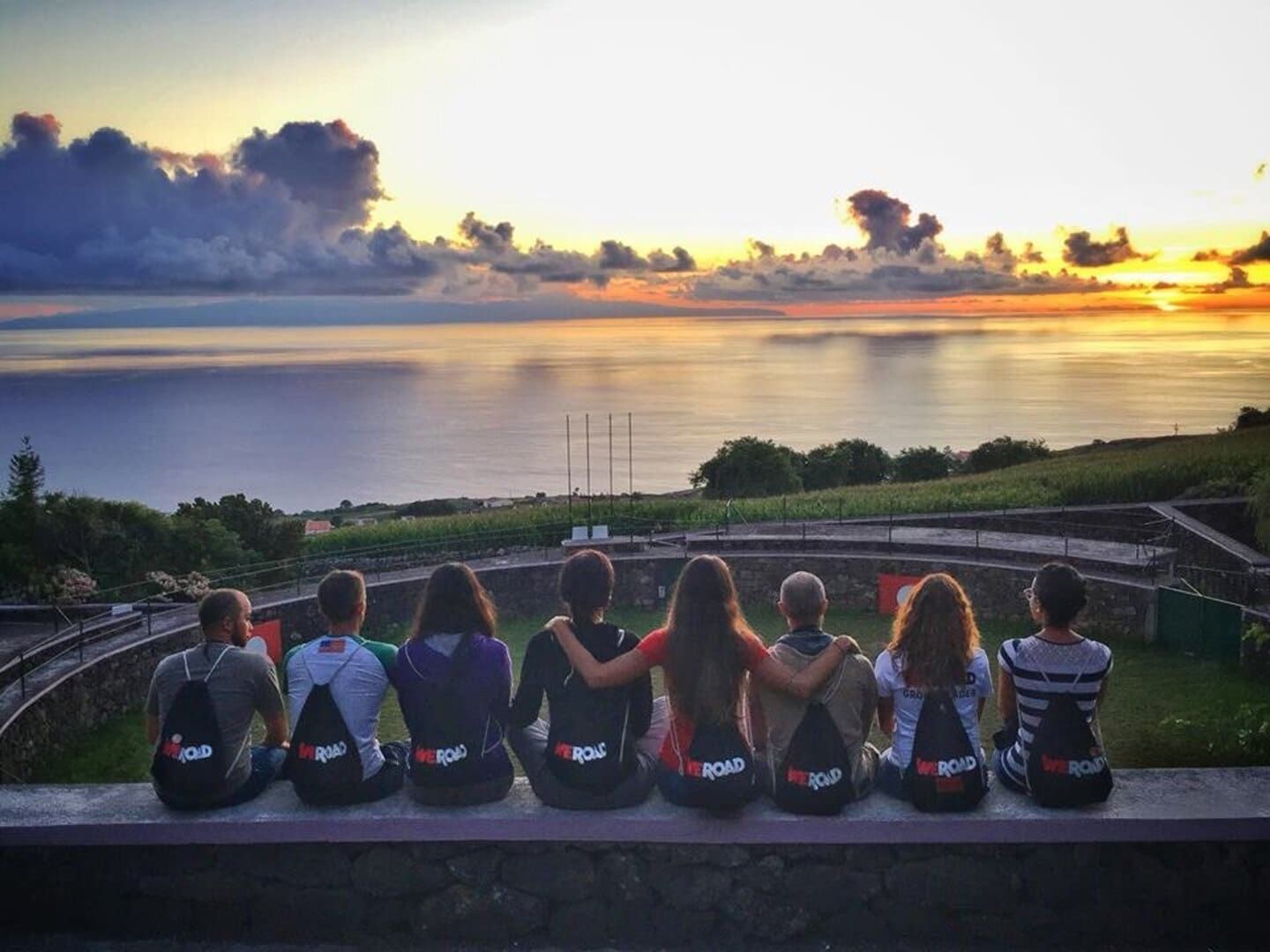 Un grupo de WeRoad se sienta en un muro de piedra, observando el atardecer sobre el océano desde un mirador verde.