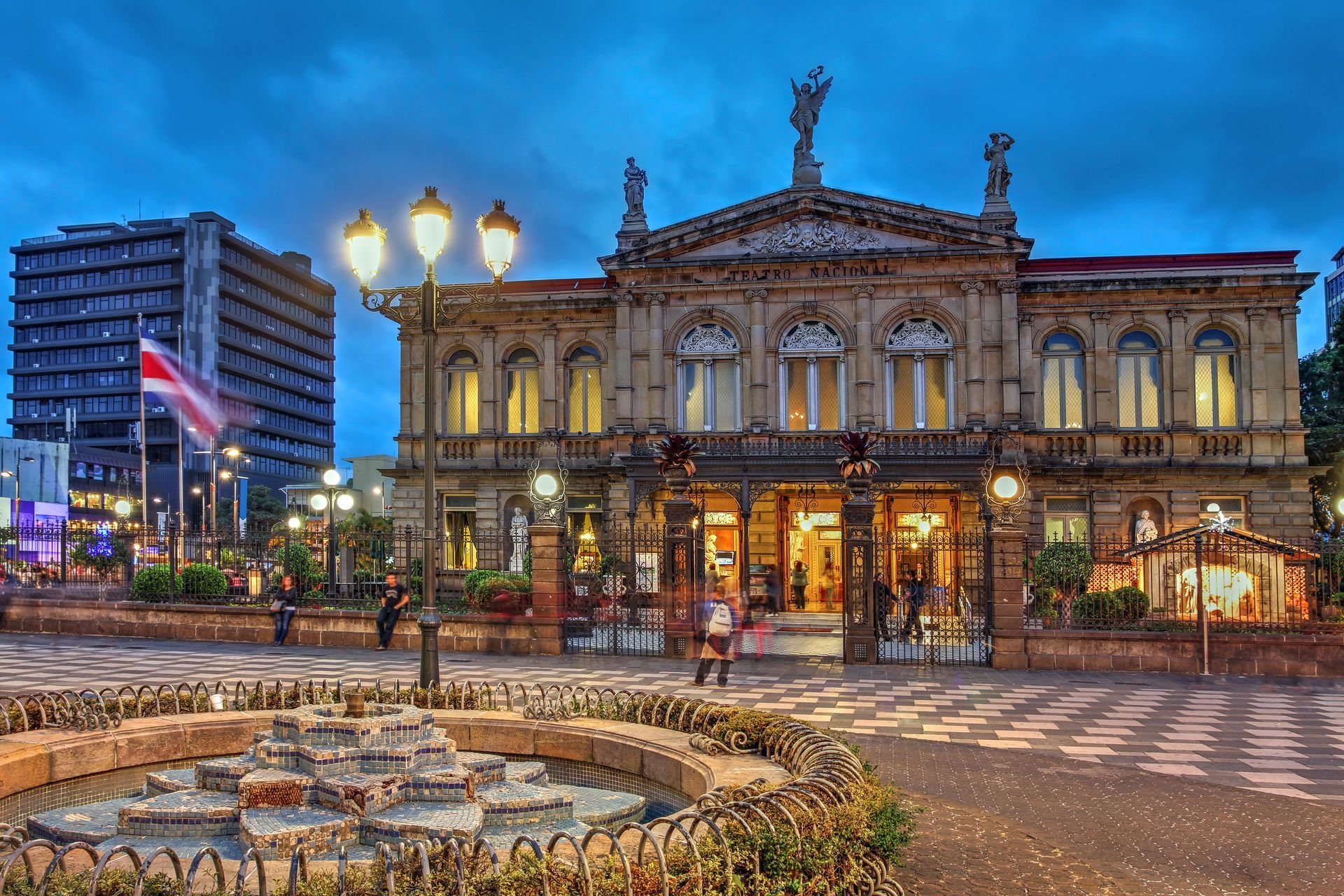 Le bâtiment orné du Théâtre National du Costa Rica, illuminé au crépuscule, avec une fontaine décorative au premier plan d'une place de ville.