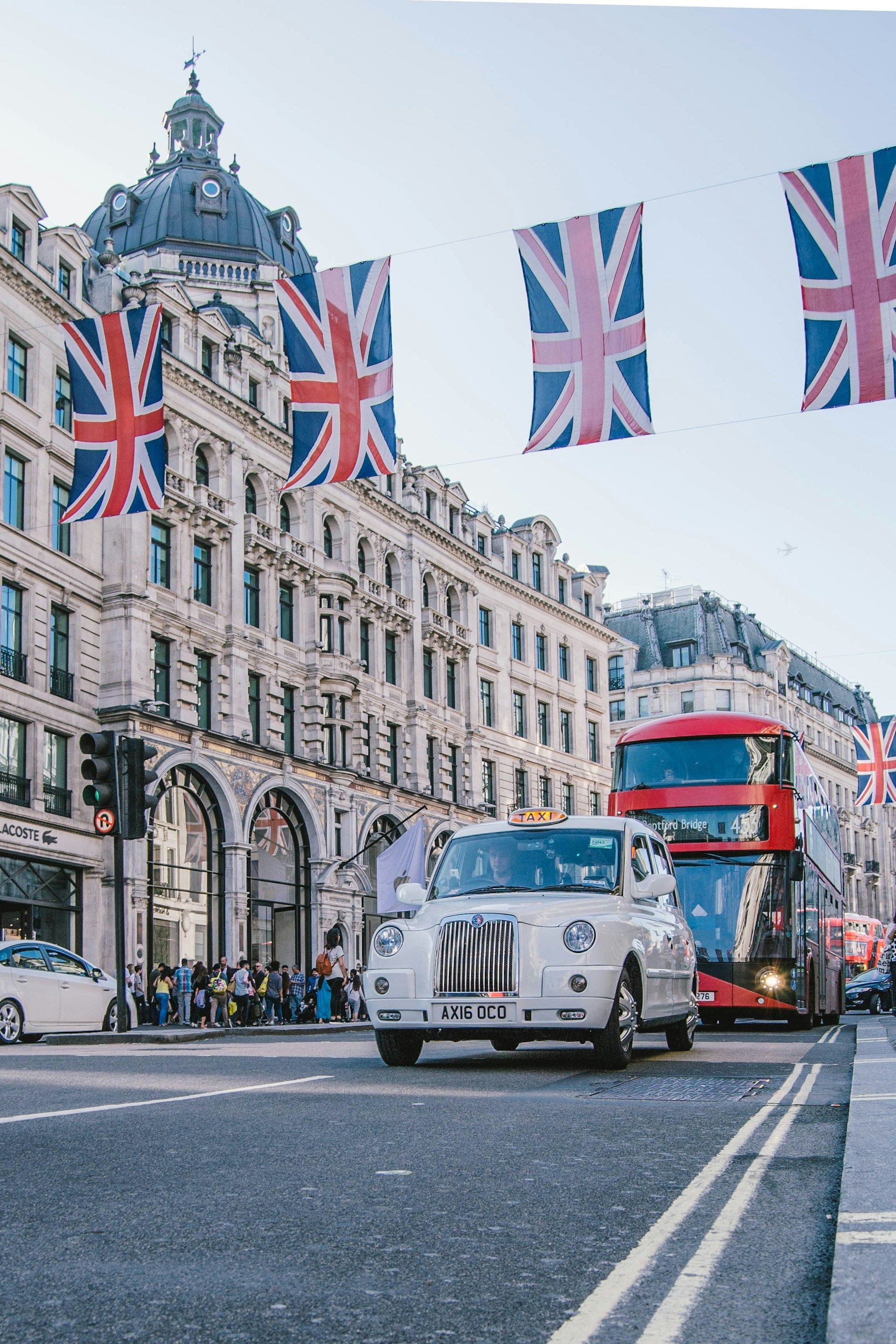 Un taxi londonien blanc et un bus à impériale rouge circulent dans une rue décorée de drapeaux Union Jack suspendus entre des bâtiments ornés.