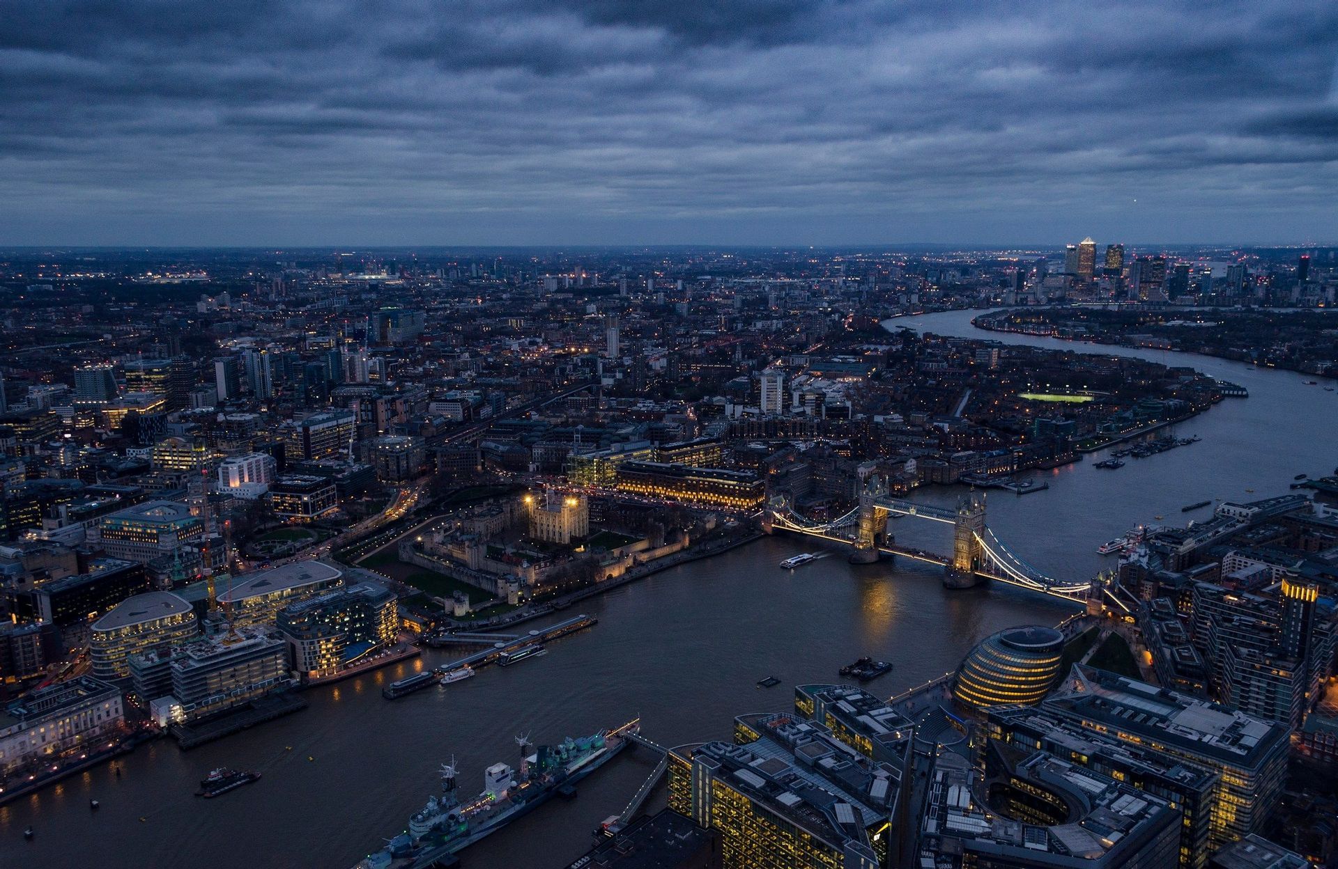 Una vista aerea del panorama urbano di Londra al tramonto, con il Tower Bridge illuminato e il Tamigi sotto un cielo nuvoloso.