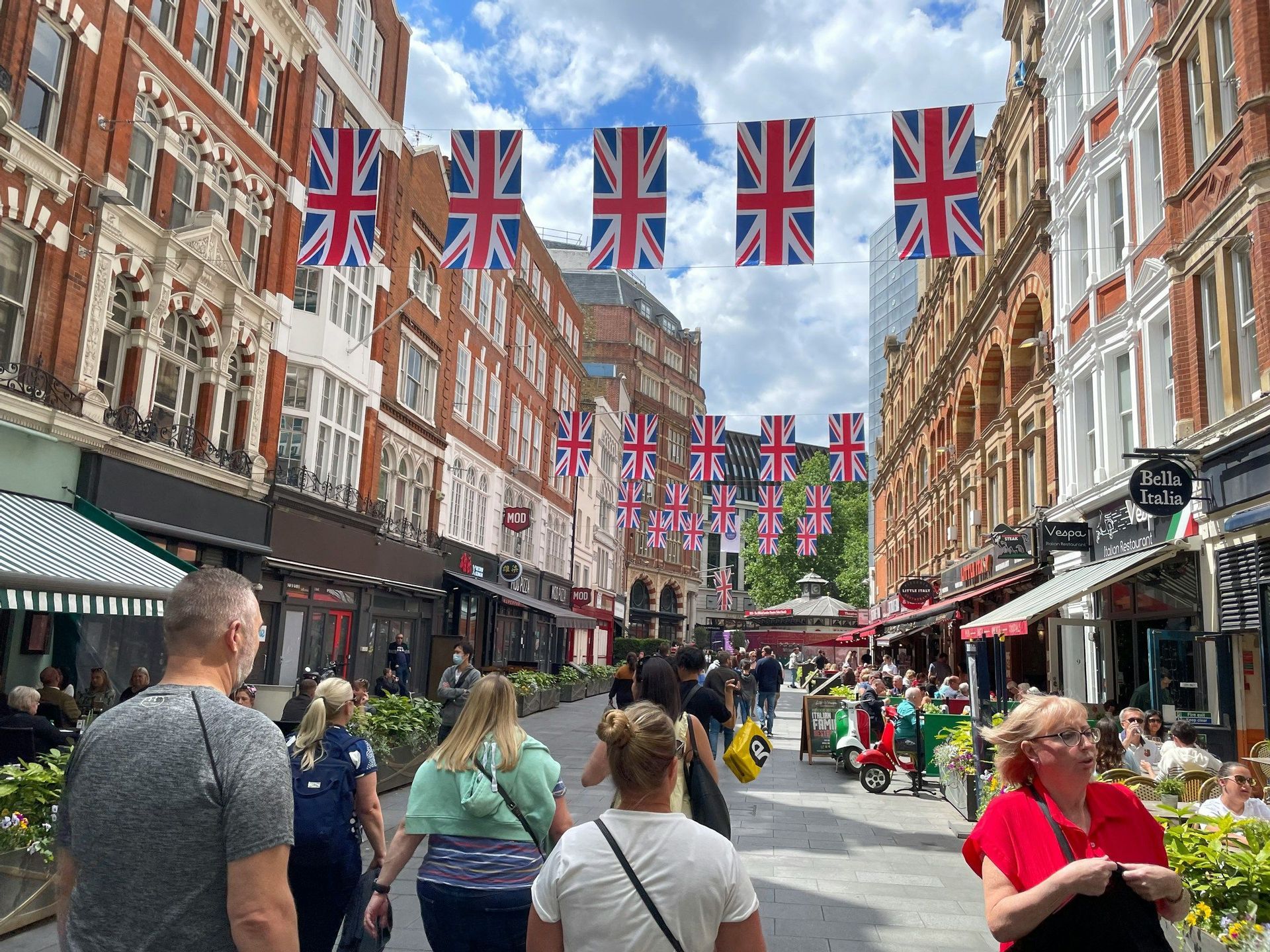 Una strada cittadina affollata con persone che camminano e cenano all'aperto, decorata con festoni di bandiere Union Jack appesi tra edifici in mattoni.