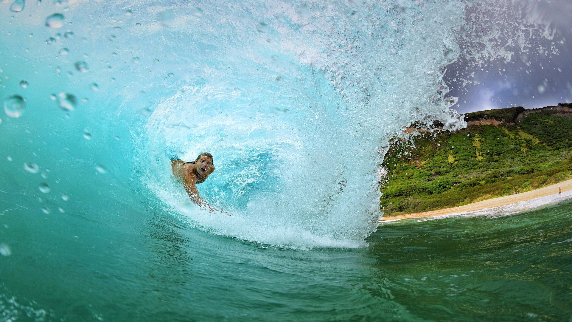 Un surfista cavalca all'interno del tubo di una grande onda turchese, con la vista che si apre verso una spiaggia sabbiosa e una costa verde.