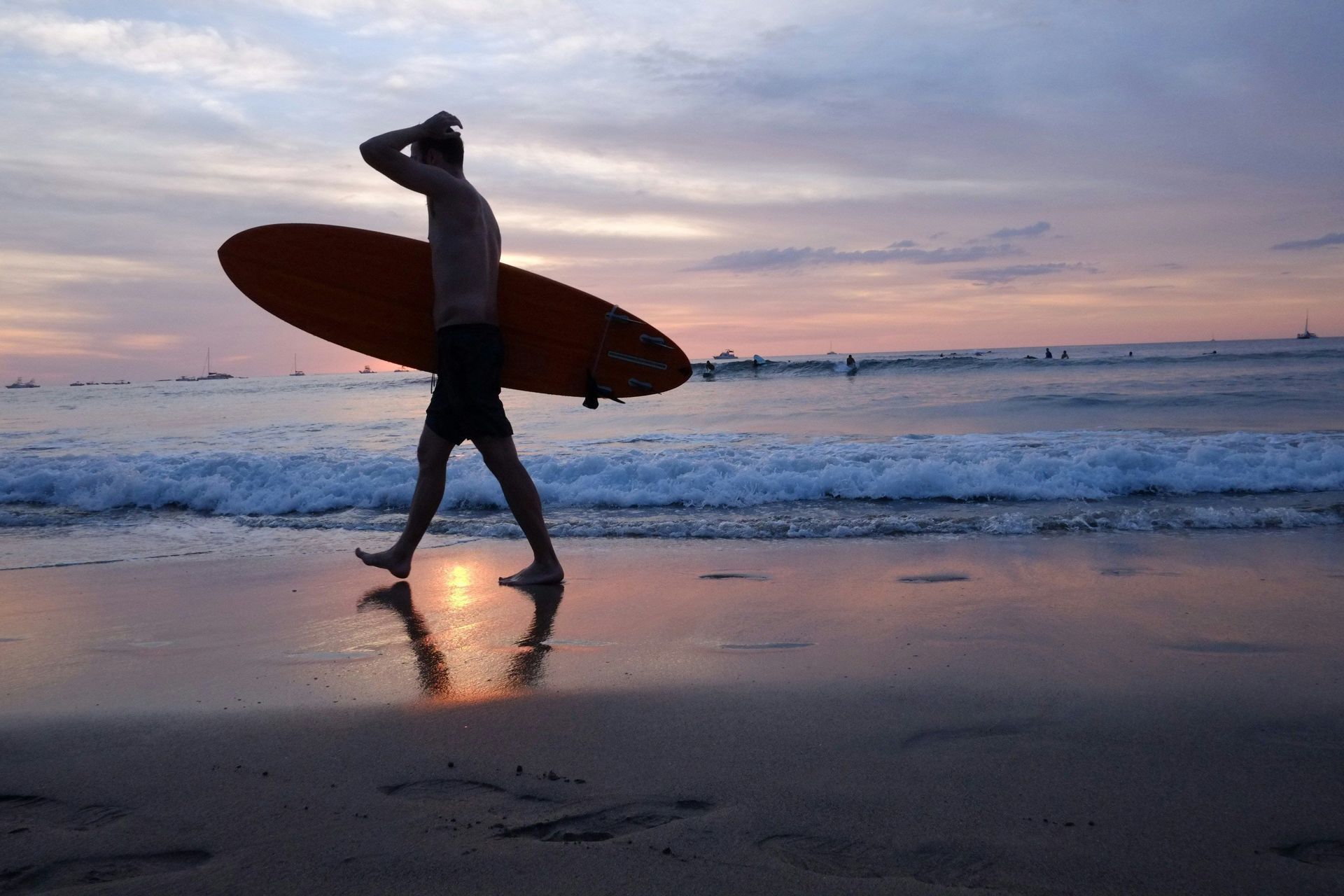 Una silhouette di un uomo con la tavola da surf cammina lungo la riva bagnata di una spiaggia durante un tramonto colorato.