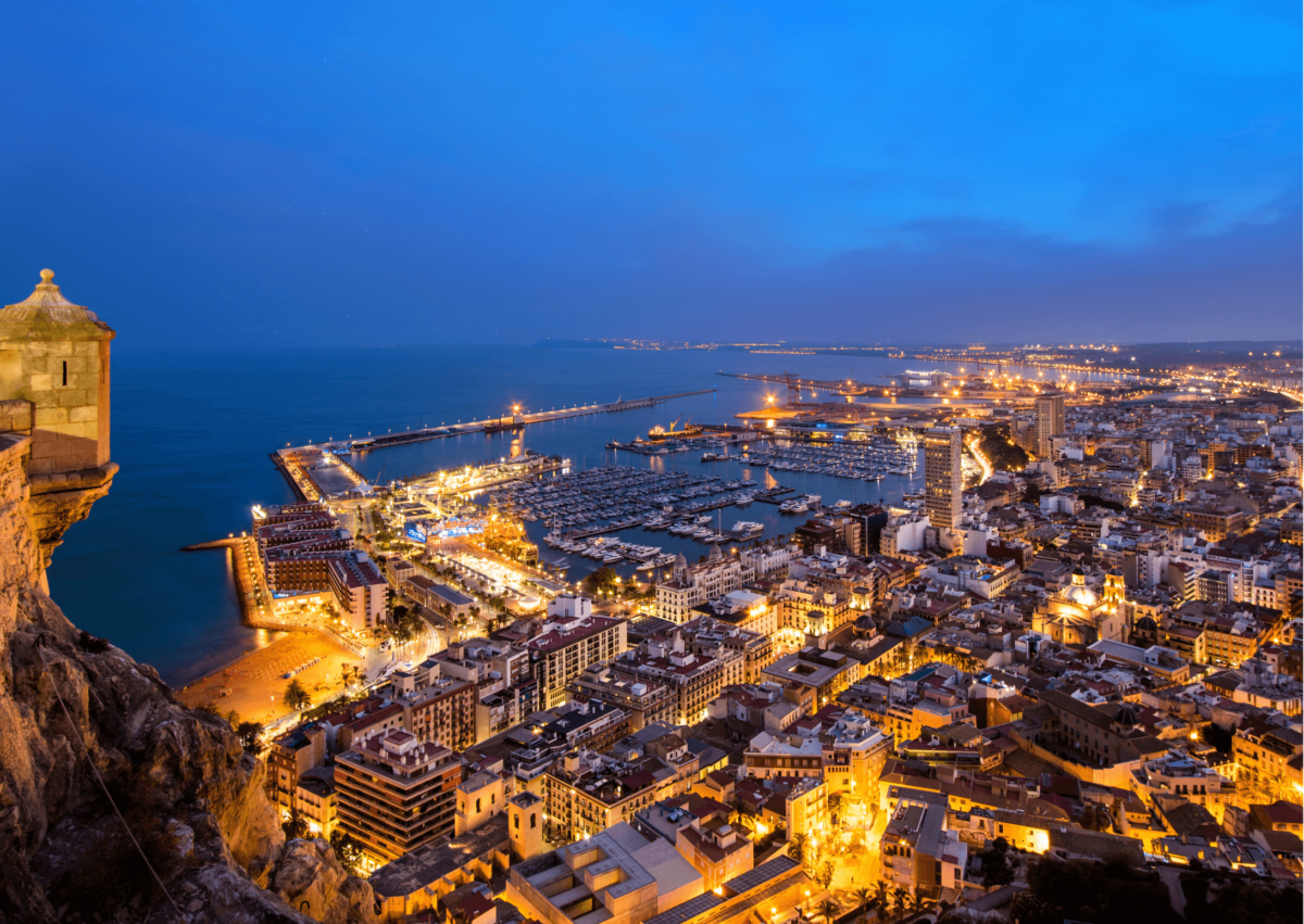 Una vista sopraelevata di una città costiera e del suo porto turistico illuminati al crepuscolo, con una torretta in pietra visibile in primo piano.