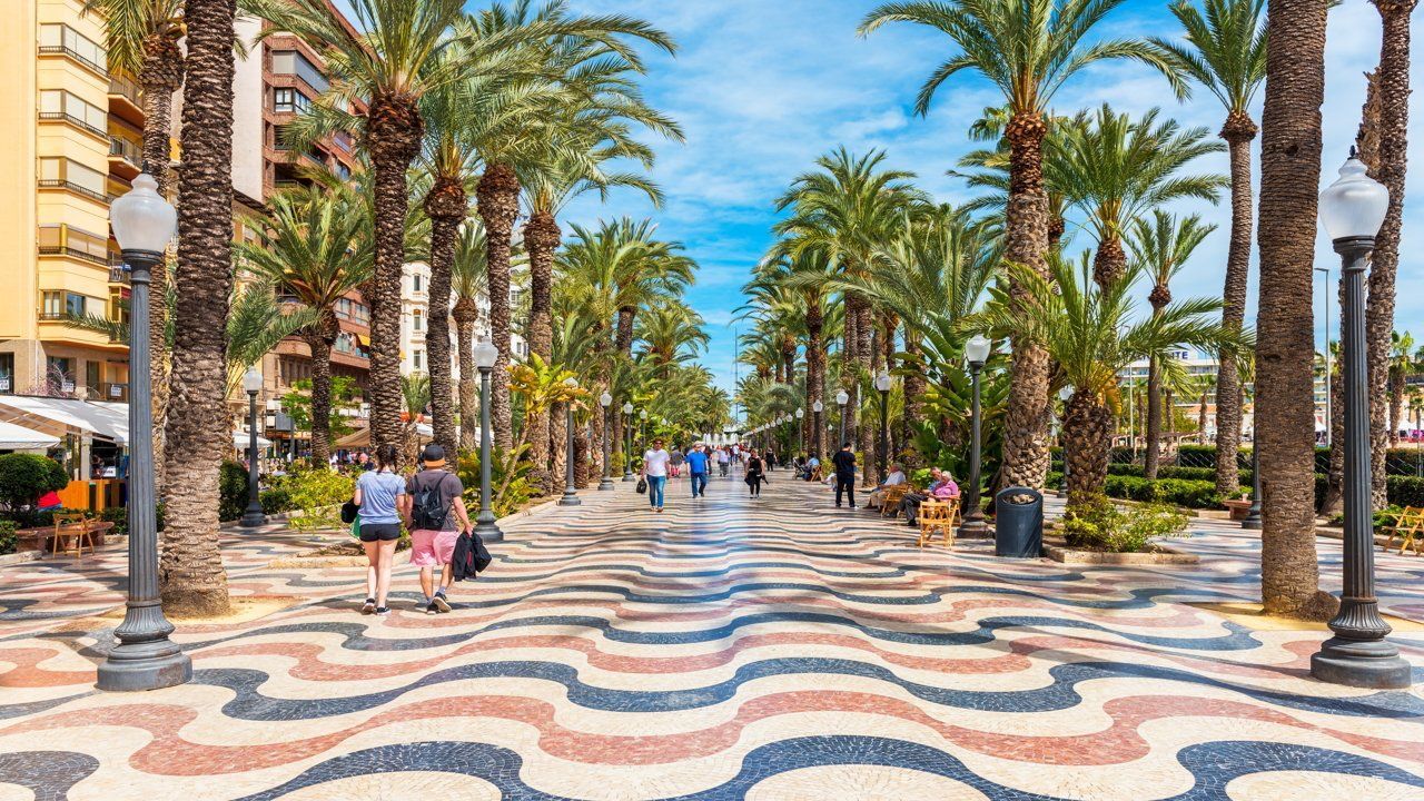 Le persone passeggiano lungo un ampio lungomare fiancheggiato da alte palme, caratterizzato da una pavimentazione a mosaico colorata e ondulata sotto un cielo azzurro.