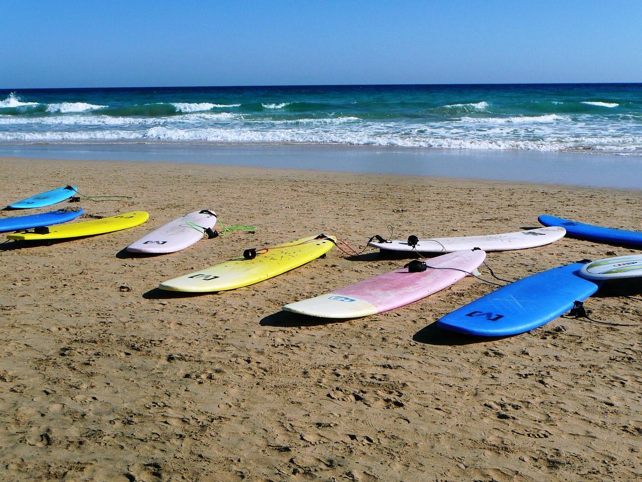 Tavole da surf colorate adagiate sulla sabbia di una spiaggia, con onde oceaniche che si infrangono sullo sfondo sotto un cielo sereno.