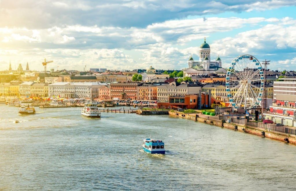 Una vista della città dall'acqua di un lungomare storico con una cattedrale, una ruota panoramica e diverse barche a vela.
