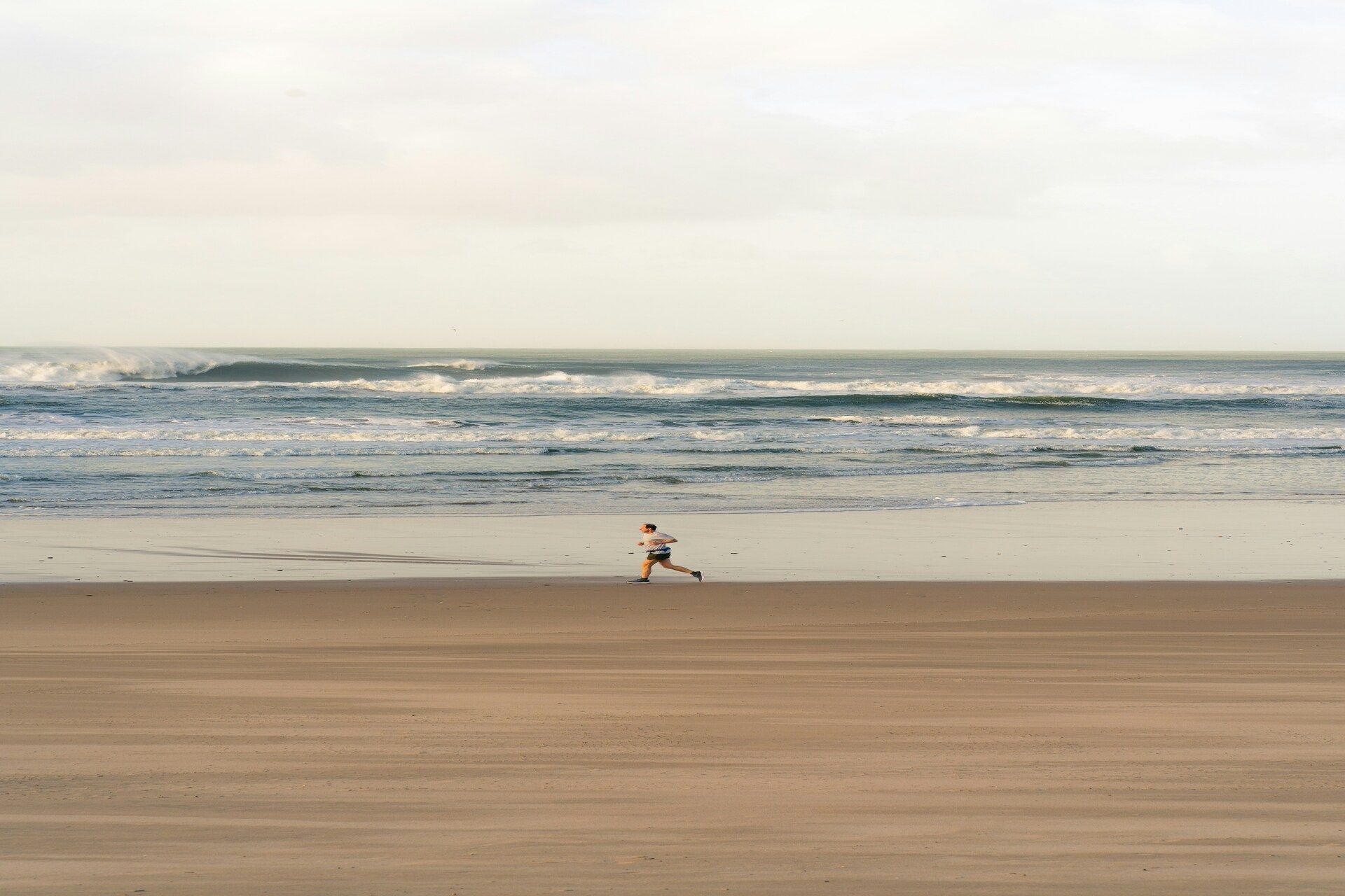 Una persona corre su una vasta spiaggia sabbiosa lungo l'oceano sotto un cielo nuvoloso.