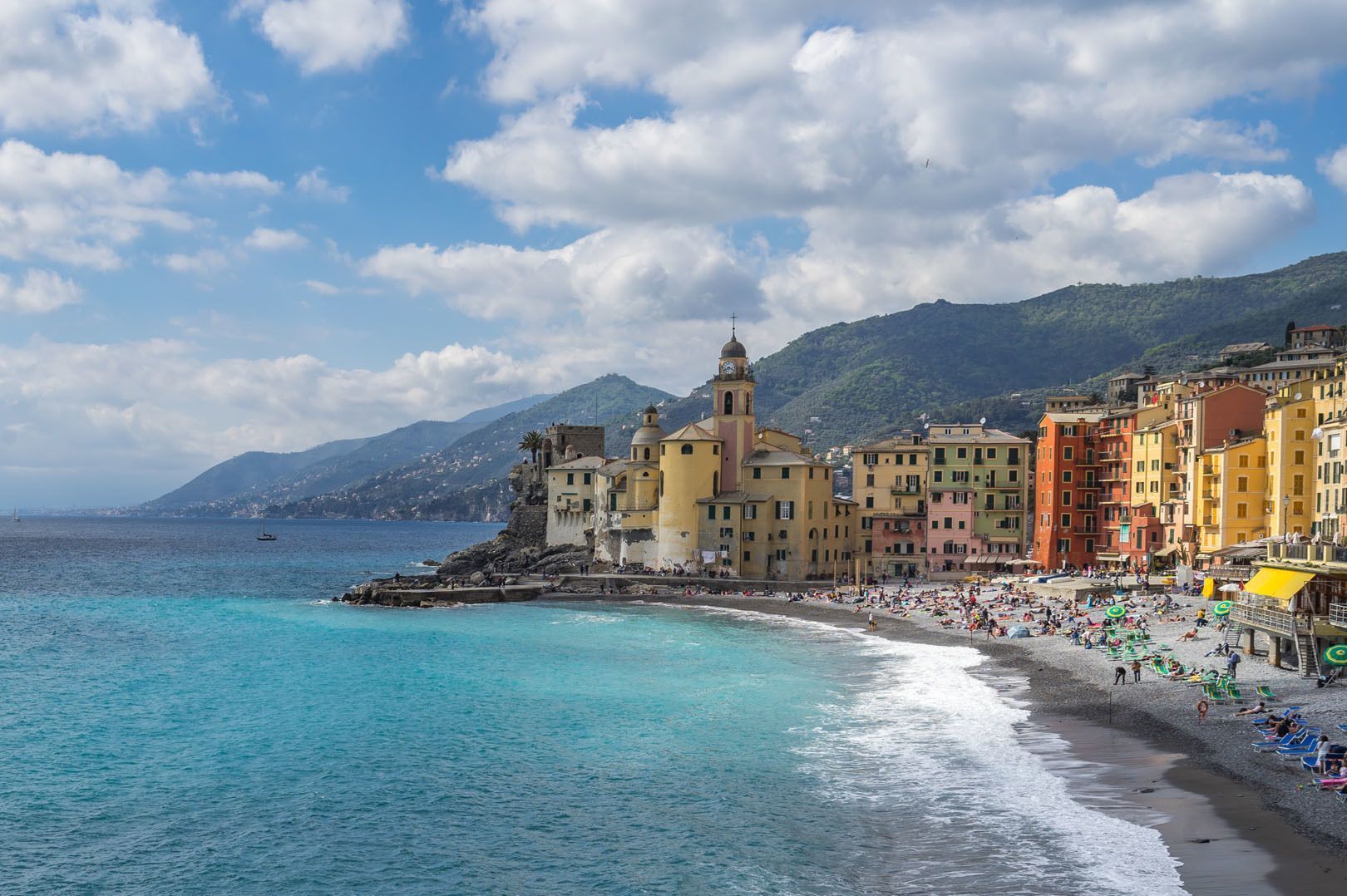 Un colorato villaggio costiero con una chiesa su una scogliera, una spiaggia di ciottoli affollata e acqua turchese.
