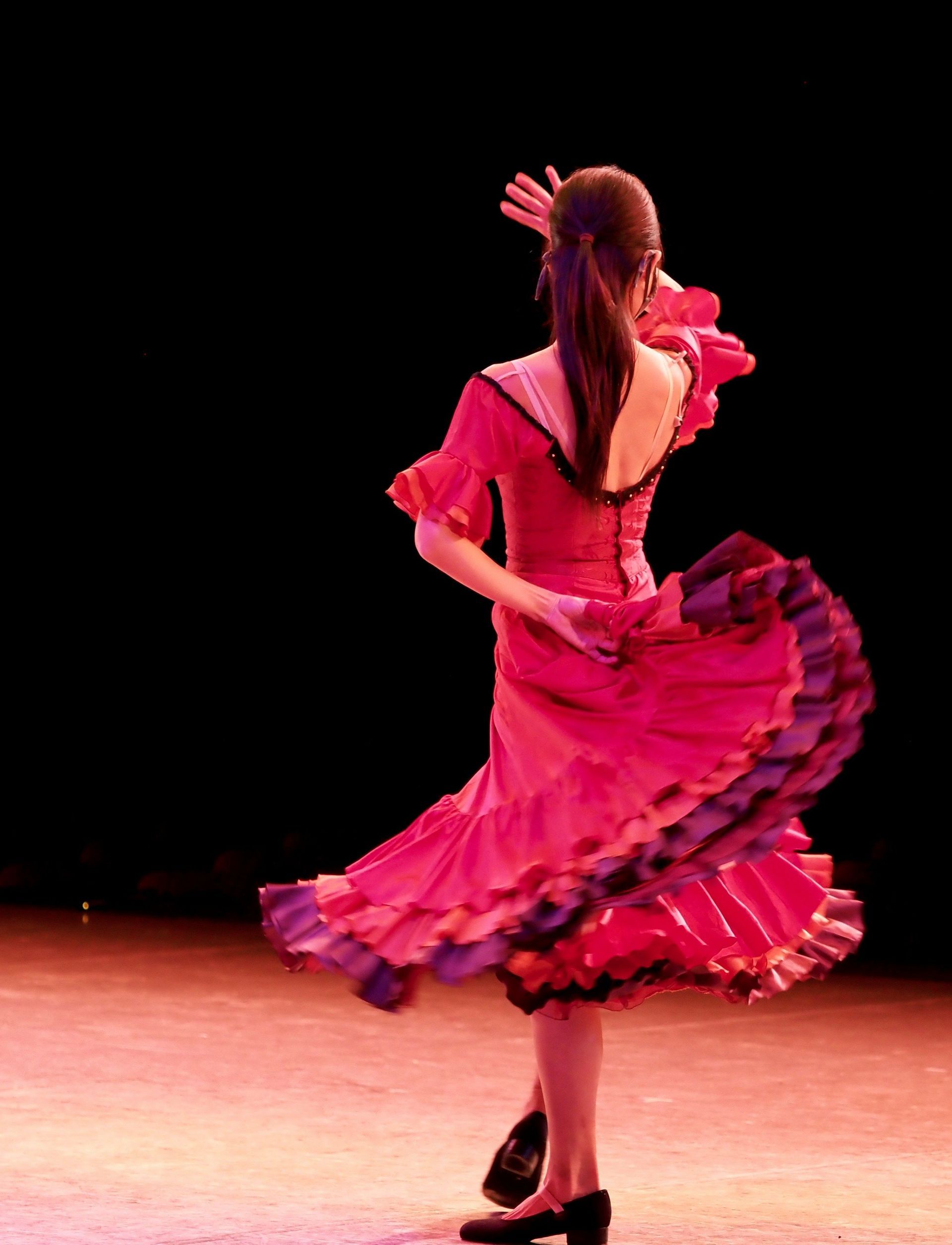 Una ballerina di flamenco, ripresa da dietro, si esibisce su un palco, il suo abito rosa e viola a balze che turbina con il movimento.