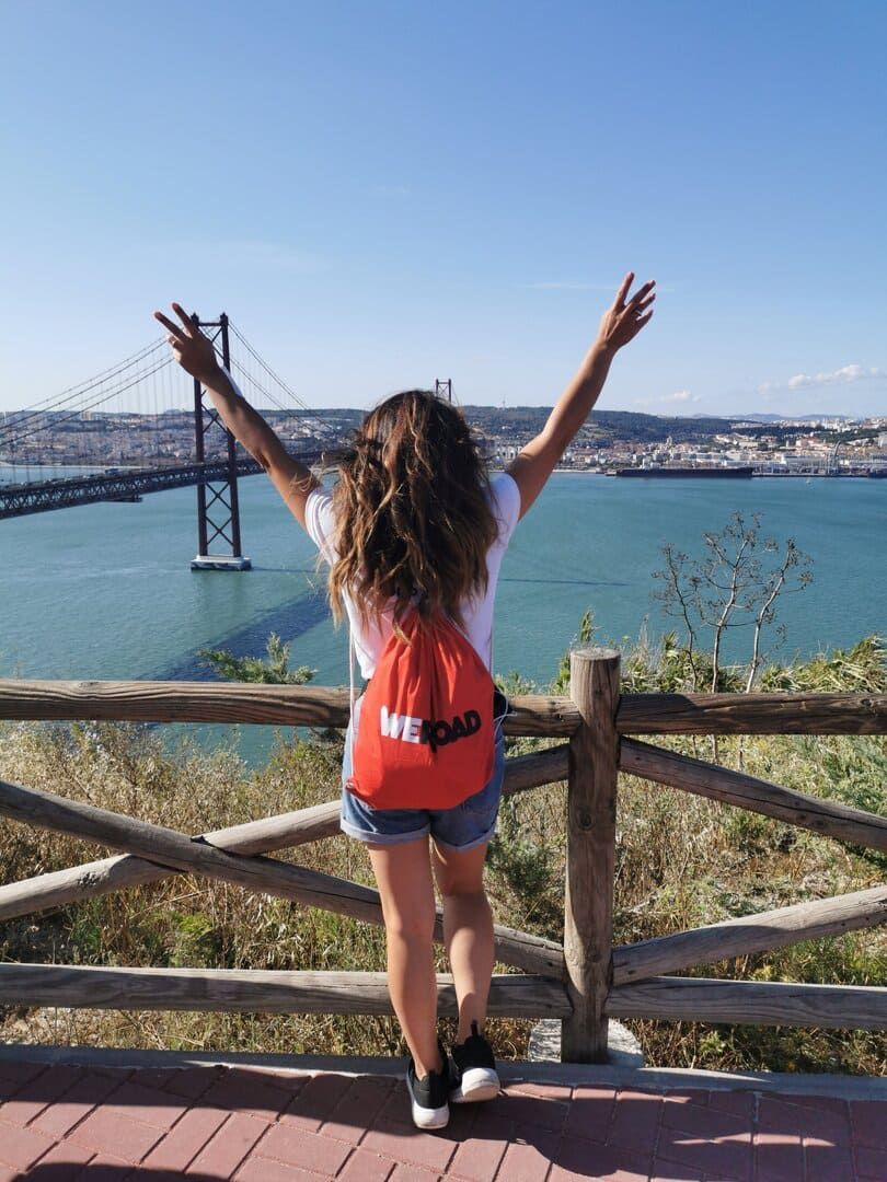 Eine von hinten gesehene Frau mit rotem WeRoad-Rucksack hebt die Arme, während sie eine große Brücke und eine Stadt am Wasser überblickt.
